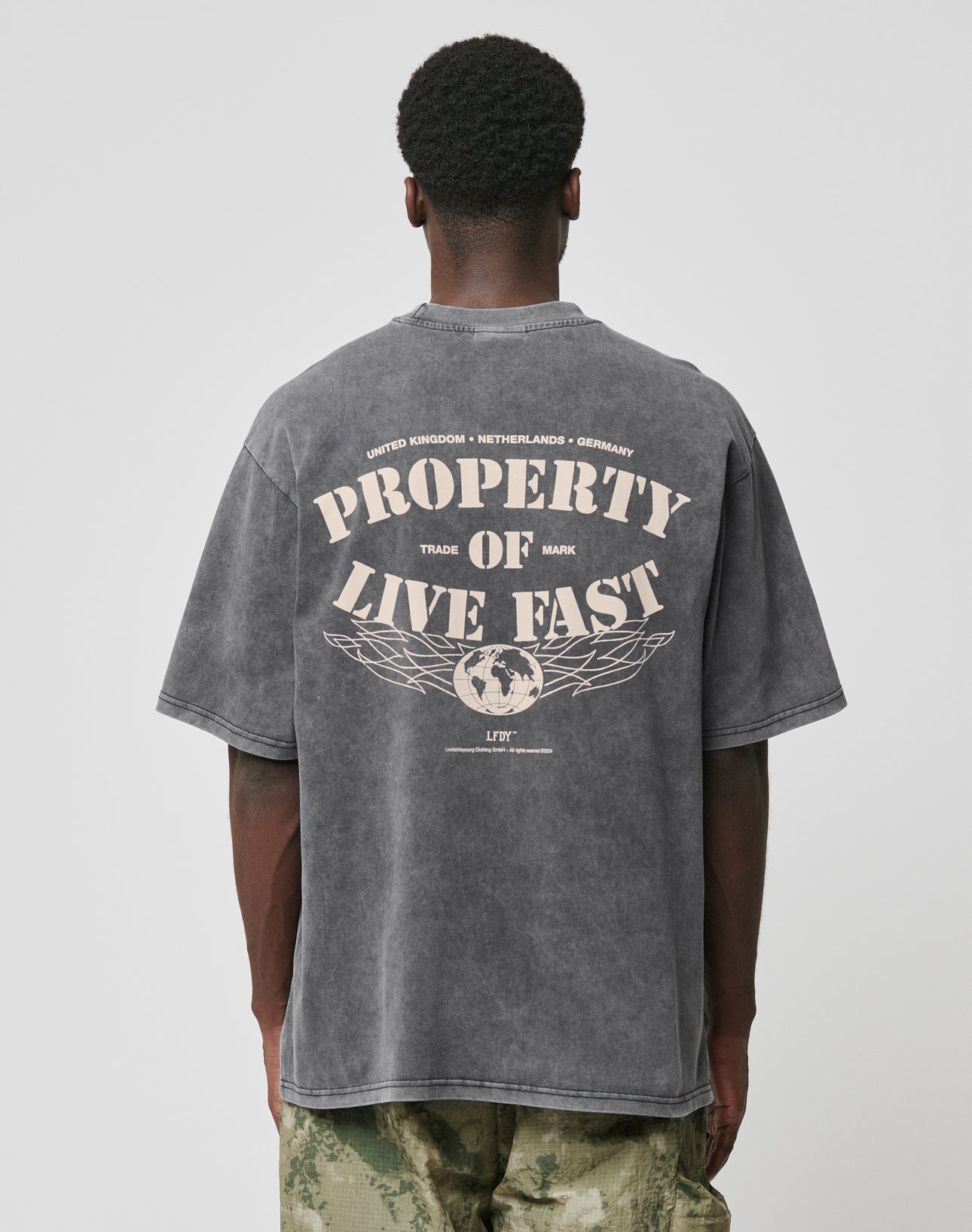 A person stands facing away, wearing the LIVE FAST DIE YOUNG LFDY Property Tee—an oversized faded black T-shirt with front and back prints (PROPERTY OF LIVE FAST and a globe graphic), paired with green camouflage pants.
