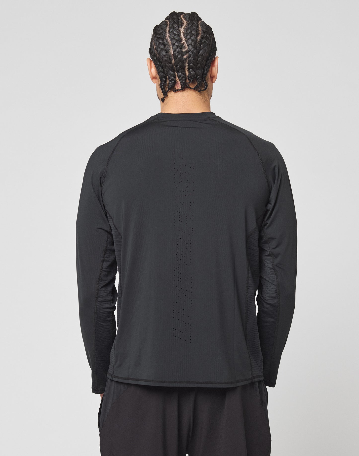 A person with braided hair is seen from behind in a LIVE FAST DIE YOUNG Live Fast Performance Longsleeve—a slim fit black athletic shirt with mesh insert and subtle perforated vertical text. The background is plain and light-colored.