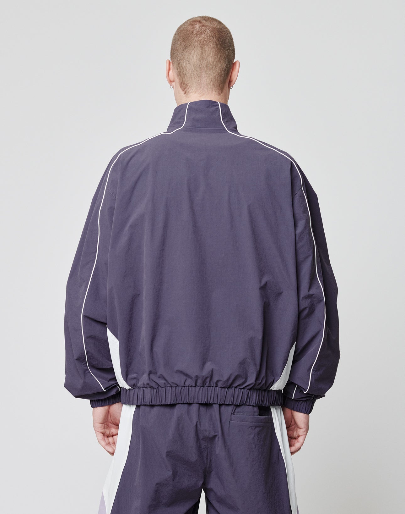 A person with short hair is shown from the back wearing the LIVE FAST DIE YOUNG Basic Nylon Trackjacket—loose-fitting, dark purple with white piping, seitliche Eingrifftaschen, and a high collar—against a plain light background.