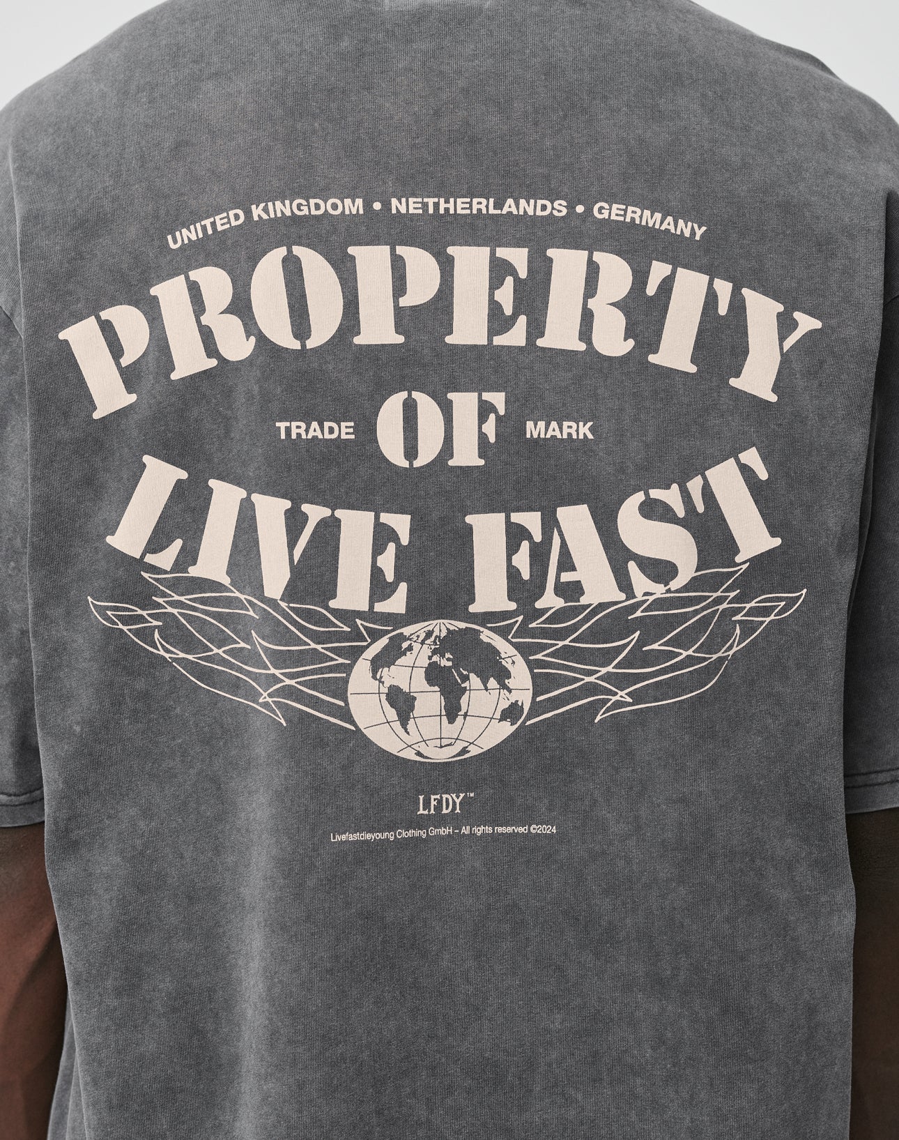 Back view of the LFDY Property Tee by LIVE FAST DIE YOUNG in faded black, featuring large tan “PROPERTY OF LIVE FAST” text above a globe and wings graphic. Front and back print; smaller text references UK, Netherlands, Germany. Oversized fit, 100% Baumwolle.