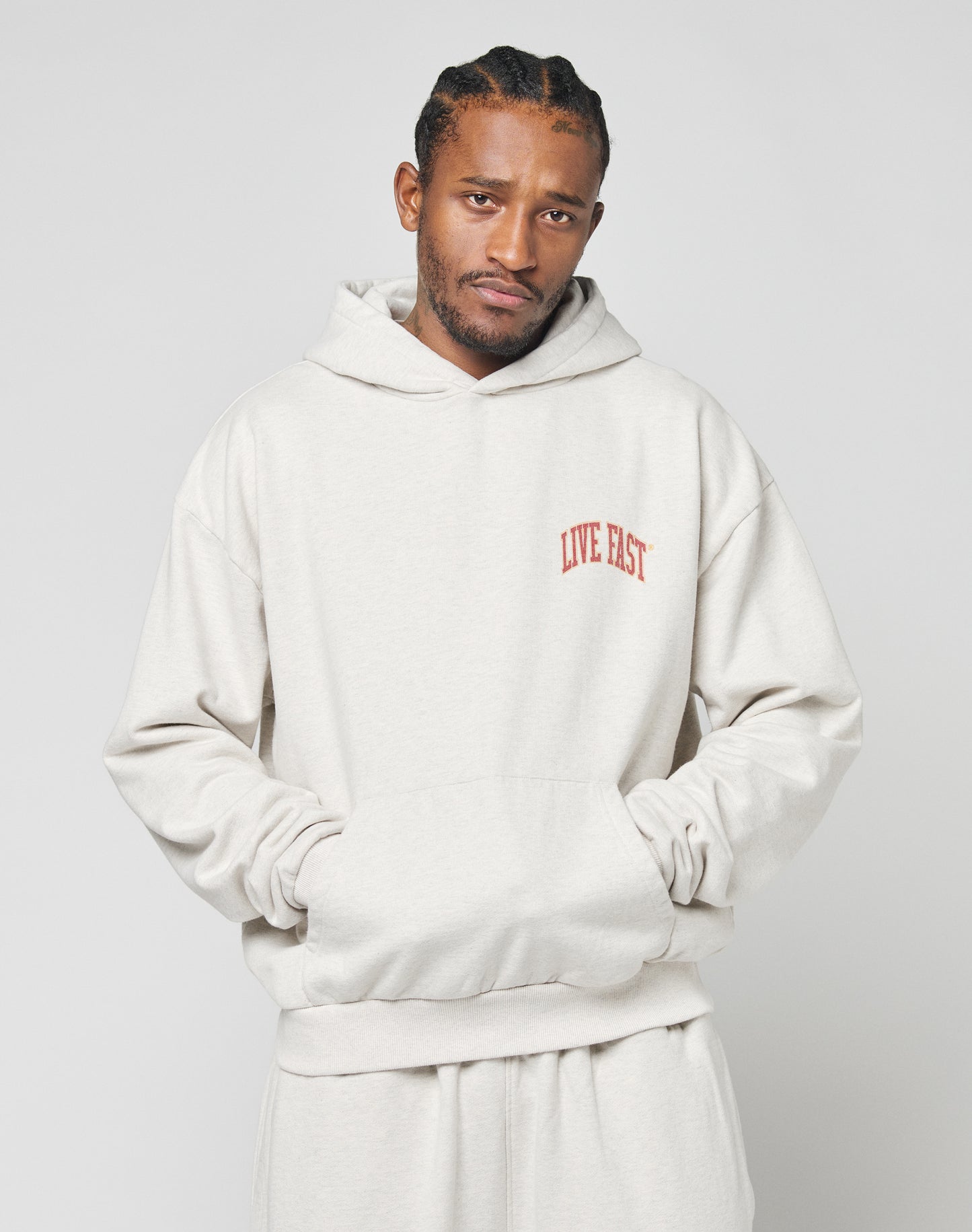 A man with braided hair wears the Live Fast Amsterdam Hooded sweatshirt and matching pants by LIVE FAST DIE YOUNG, in oversize fit light beige 100% Baumwolle, featuring a red LIVE FAST chest print. He stands hands in pockets against a plain background.