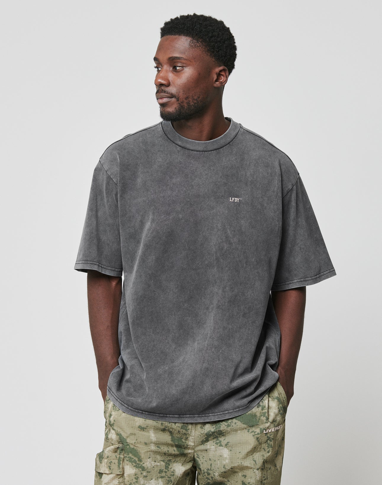 A man wears the LIVE FAST DIE YOUNG LFDY Property Tee, an oversized faded gray 100% Baumwolle shirt with small white chest text, paired with green camo pants. He stands against a plain light gray background, looking left with hands in pockets.