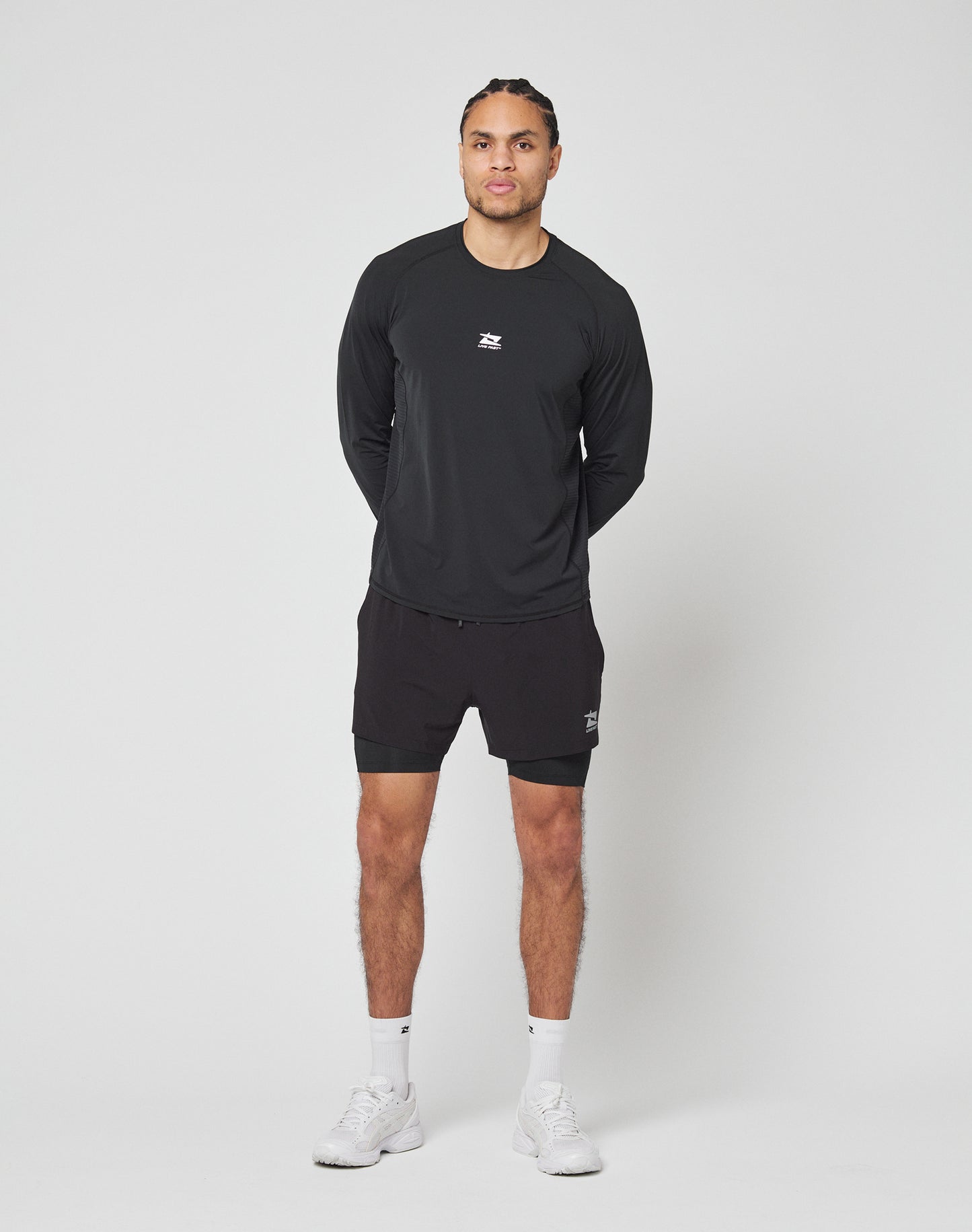 A person stands against a plain white background, wearing LIVE FAST DIE YOUNGs black Live Fast Performance Running Shorts with integrated tights, a black long-sleeve athletic shirt, white socks, and white sneakers. Hands behind back, facing forward.