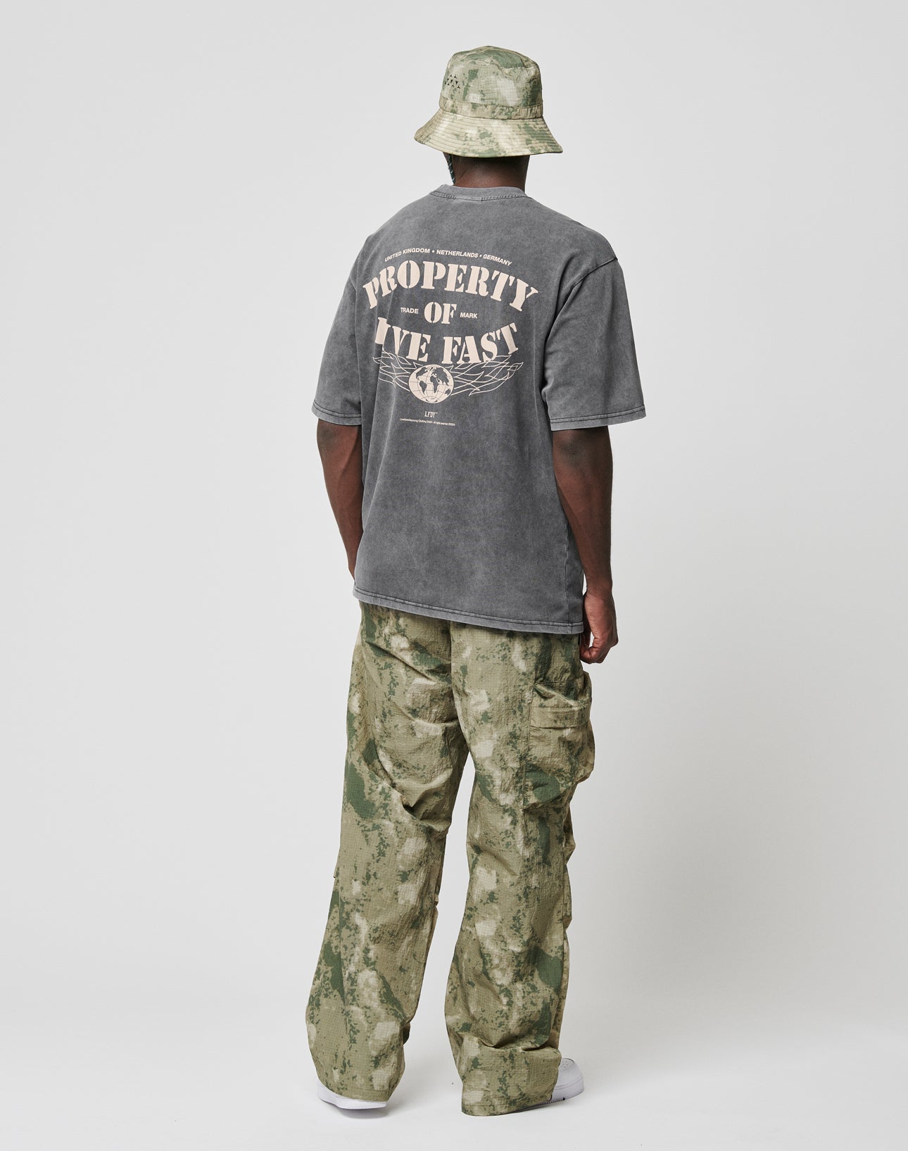 A person in an oversized gray LIVE FAST DIE YOUNG LFDY Property Tee with front and back print, loose camo pants, white sneakers, and a camo bucket hat stands facing away against a plain white background.
