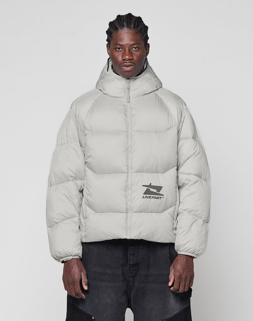 A man with braided hair wears the LIVE FAST DIE YOUNG Live Fast Puffer Jacket, a light gray, oversized, hooded style featuring a reflektierender Print and LIVE FAST graphic, standing against a plain white background.