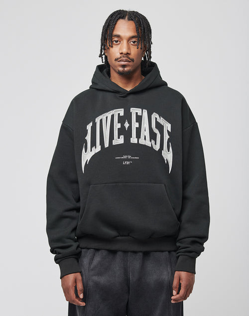 A man with short dreadlocks wears the LIVE FAST DIE YOUNG Live Fast Axe Hooded, an oversize black Baumwolle-Polyester hoodie with bold white LIVE FAST text, standing against a plain light gray background.