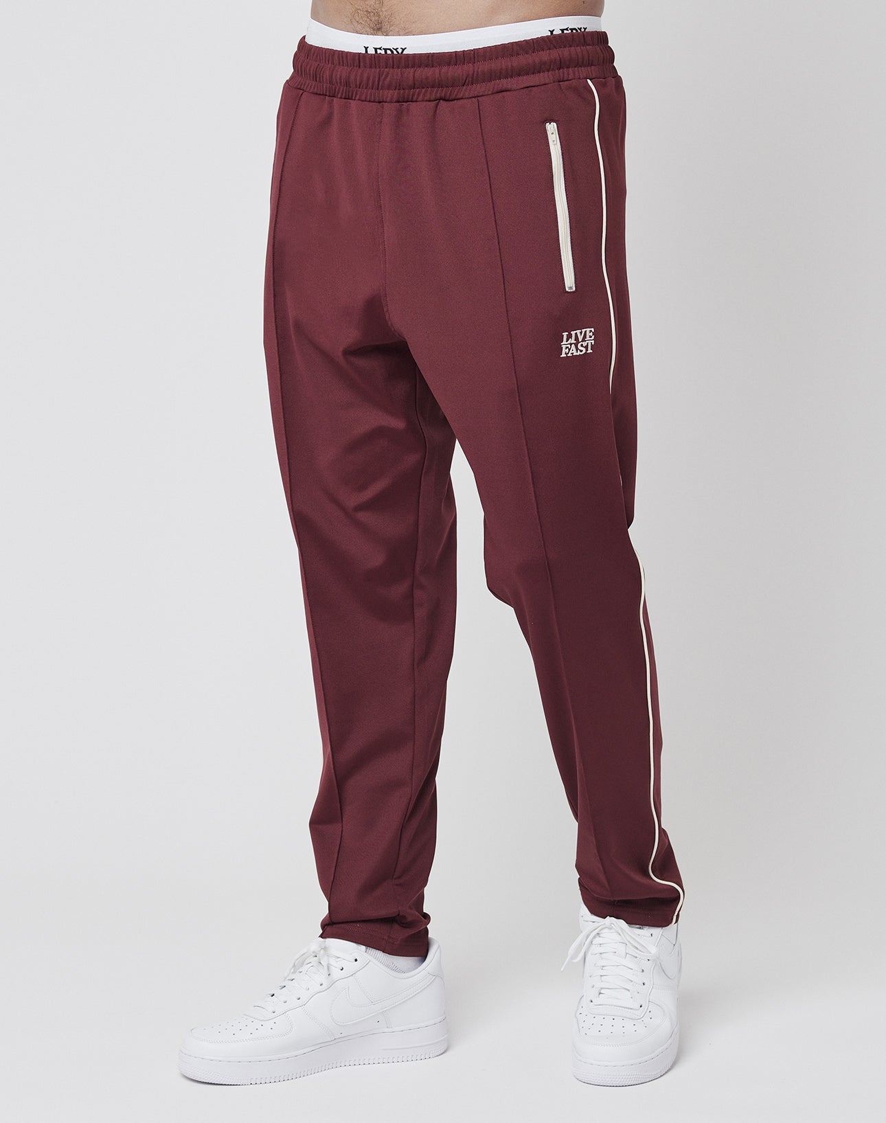 A person wears the LIVE FAST DIE YOUNG Live Fast Trackjogger in maroon with white side stripes, zipper pockets, and LIVE FAST text on the upper left thigh, paired with white sneakers against a plain white background.