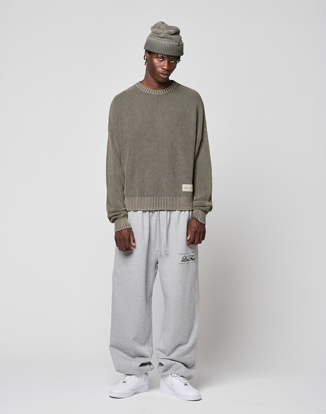 Wearing the LFDY Knit Sweater by LIVE FAST DIE YOUNG in olive green with a dropped shoulder and oversized fit, plus a matching knit beanie, loose light gray sweatpants with lettering, and white sneakers, this person stands against a plain light gray background.