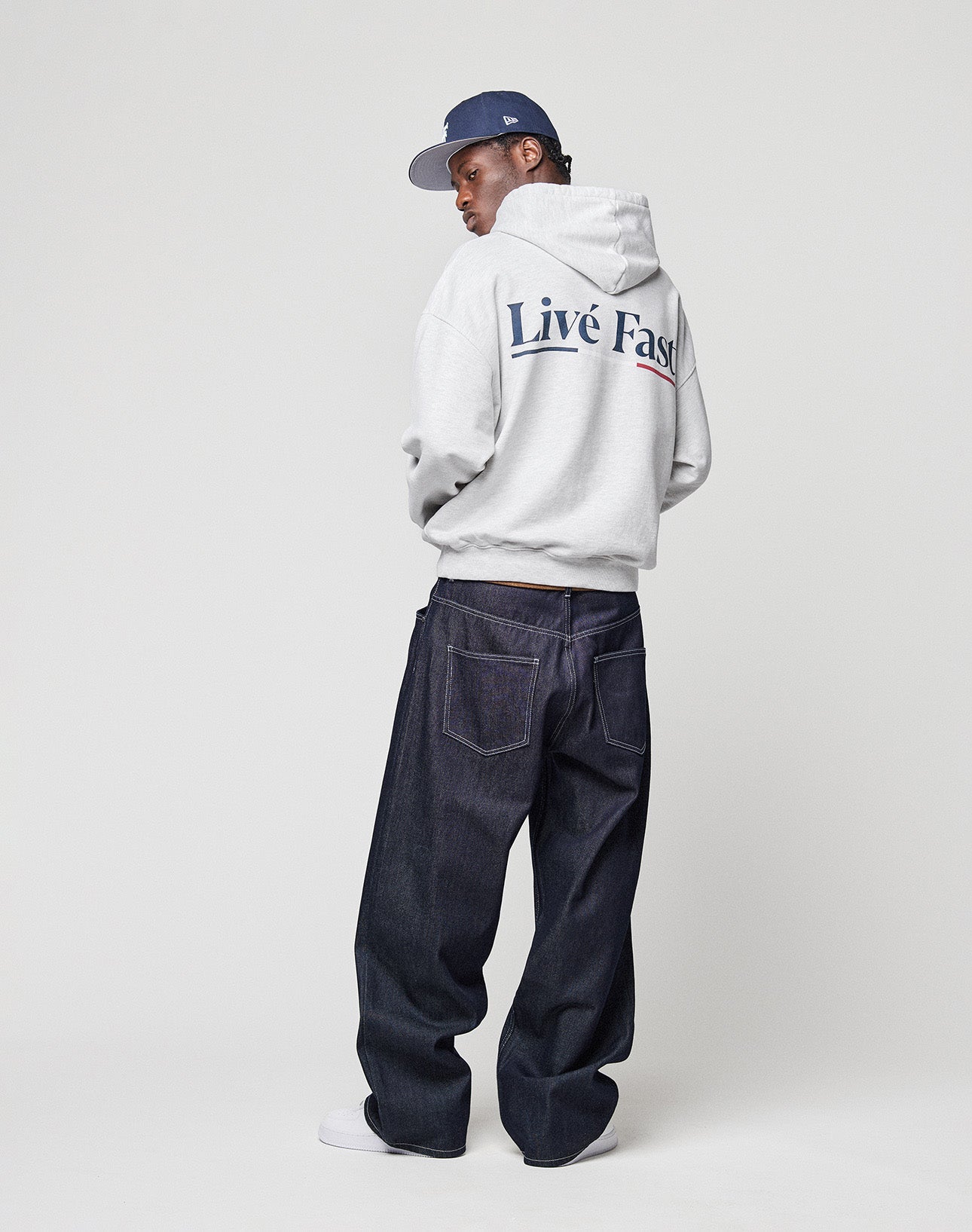 A man wears an oversized gray Live Fast Paris Hooded by LIVE FAST DIE YOUNG with front and back print, paired with dark baggy jeans, white sneakers, and a blue cap in a minimalist studio.