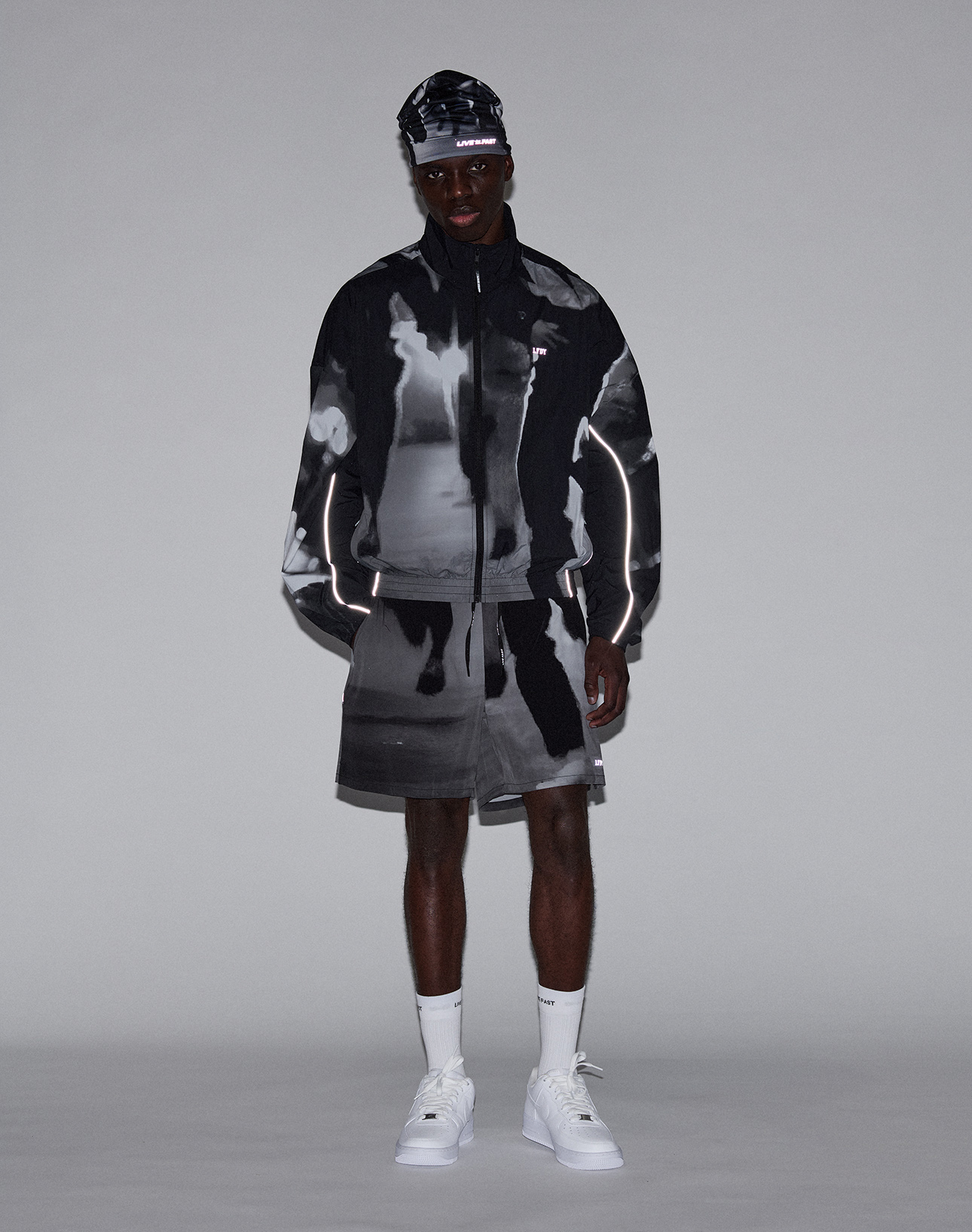 A man poses against a plain background wearing the LIVE FAST DIE YOUNG LFDY Performance Jacket with matching shorts, white socks and sneakers, and a coordinating cap. The outfit features an allover black and white print with reflective logo sleeves.