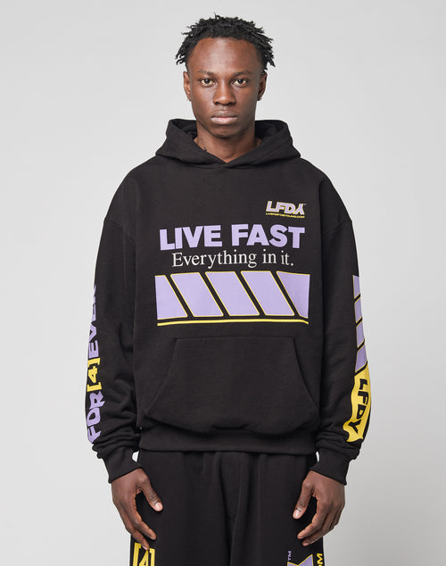 A person in an oversize fit black LFDY Racing Hooded by LIVE FAST DIE YOUNG, featuring glossy white and lavender LIVE FAST Everything print with yellow and lavender stripes, plus matching black pants, stands against a light gray background.