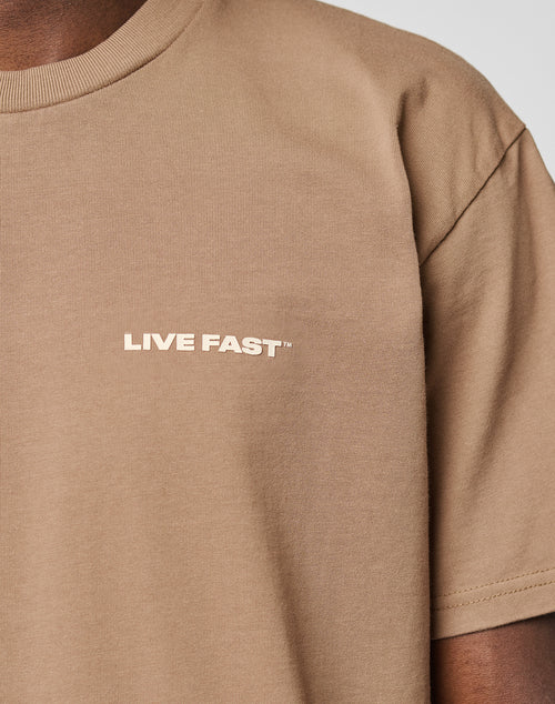 Close-up of a person wearing the beige Live Fast Plain Tee by LIVE FAST DIE YOUNG, featuring a bold LIVE FAST front print. Only the upper torso and part of the left arm are visible. Made from 100% Baumwolle in regular fit.