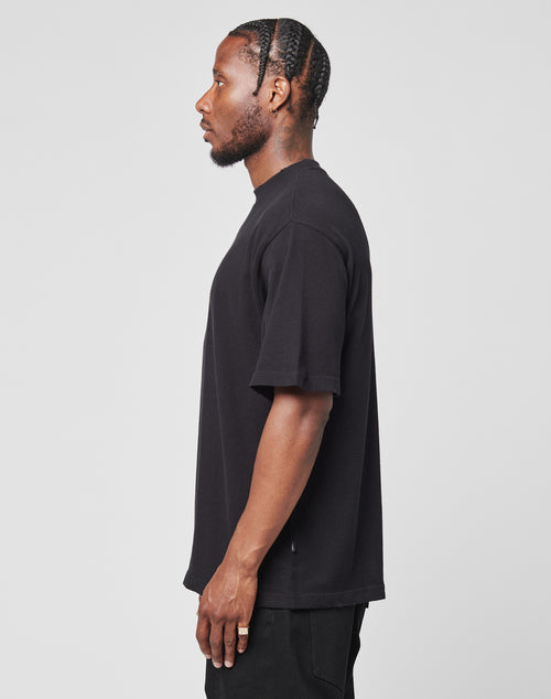 A man with braided hair stands in profile against a plain background, wearing the LIVE FAST DIE YOUNG LFDY Vintage Tee—an oversized black t-shirt with distressed edges—paired with black pants.