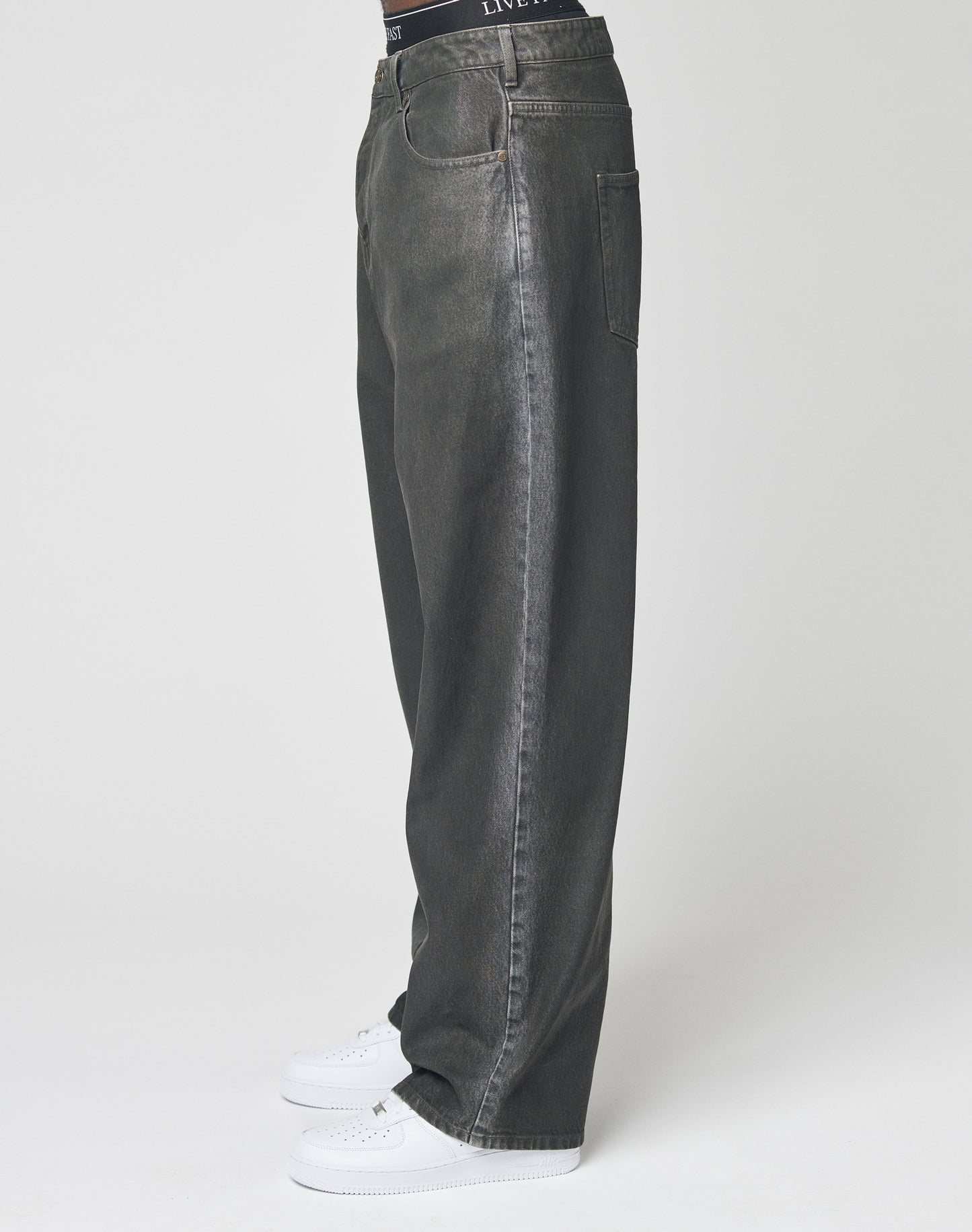 A person wearing LIVE FAST DIE YOUNG’s LFDY Coated Baggy Denim with straight legs and white sneakers stands against a plain white background, showing only the lower half of the body in side profile.