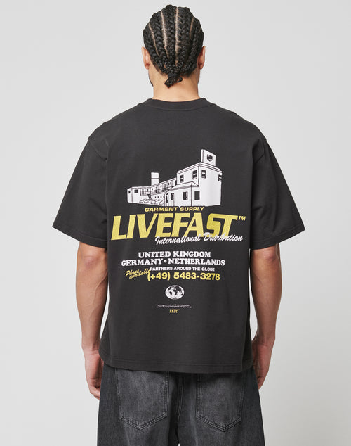 A person with braided hair wears the oversized fit LFDY Supply Tee by LIVE FAST DIE YOUNG, featuring a frontprint and a backprint with a white building graphic, yellow “LIVE FAST” text, international locations, and a phone number.