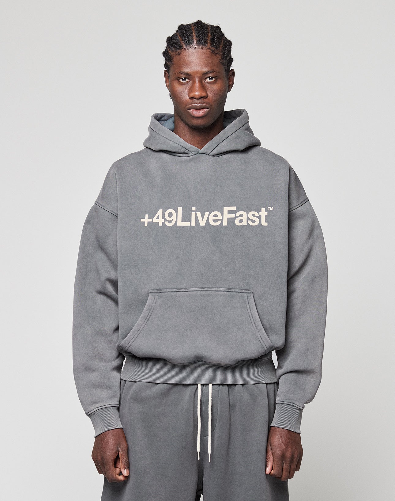 A man faces the camera in a gray LIVE FAST DIE YOUNG Basic +49 Hooded sweatshirt and matching sweatpants, featuring a bold white "+49LiveFast" front print. The background is plain and light-colored.