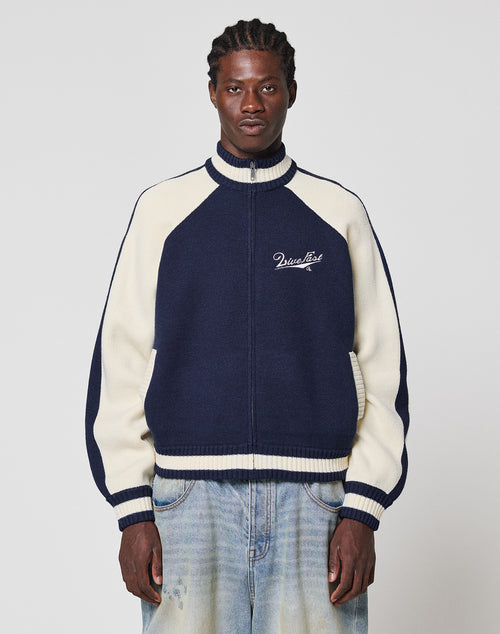 A man with braided hair wears a navy blue and cream Boxy Knit Zip Jacket by LIVE FAST DIE YOUNG, paired with light blue jeans, standing against a plain light background.