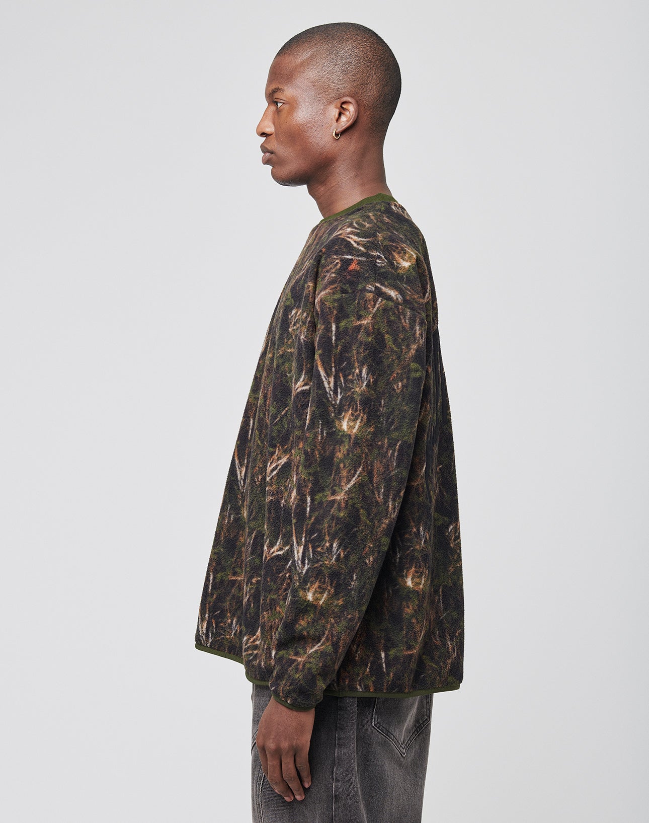 A person stands in profile against a plain background, wearing the LFDY Fleece Longsleeve by LIVE FAST DIE YOUNGāa polyester, long-sleeved, oversize-fit shirt with a camouflage patternāpaired with dark jeans.