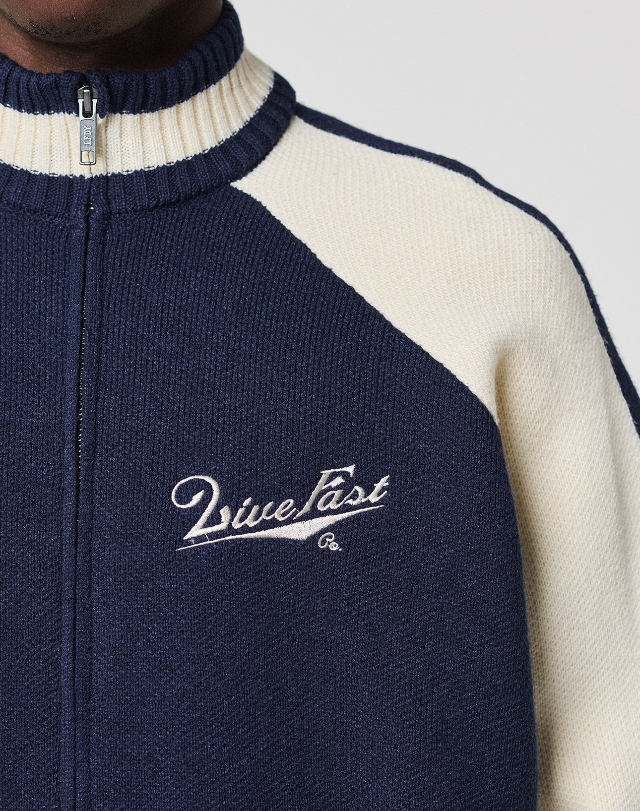 A close-up shows a person in a navy blue and cream Boxy Knit Zip Jacket by LIVE FAST DIE YOUNG. "Live Fast" is embroidered in white on the left chest. Only the neck, shoulder, upper chest, and raglan sleeves are visible.