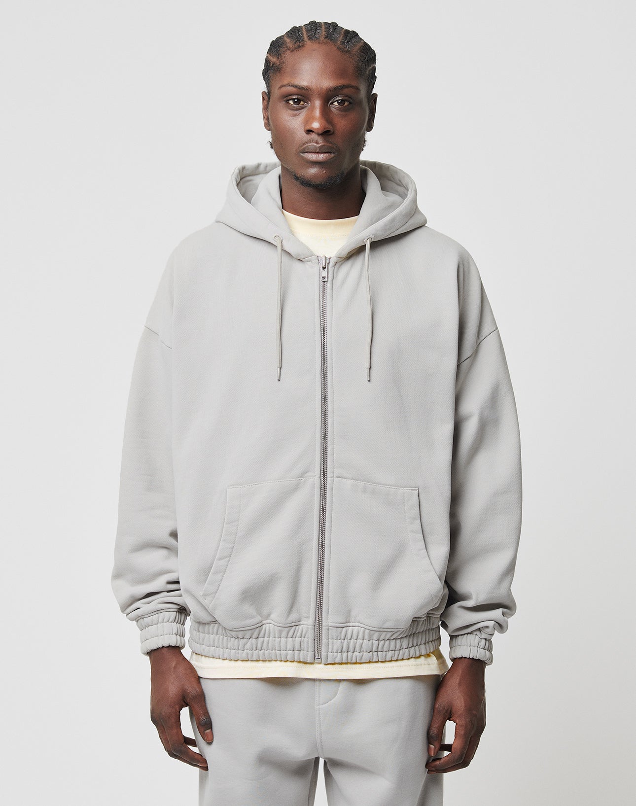 A person with braided hair faces forward, wearing the LIVE FAST DIE YOUNG Basic 510 Hooded Zip in light gray over a cream shirt and matching gray pants, set against a plain white background.