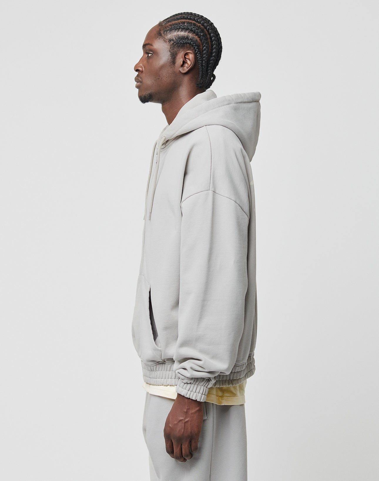 A man with braided hair stands in profile against a white background, wearing the LIVE FAST DIE YOUNG Basic 510 Hooded Zip and matching sweatpants in light gray, both made of soft Baumwolle. His hands rest at his sides.