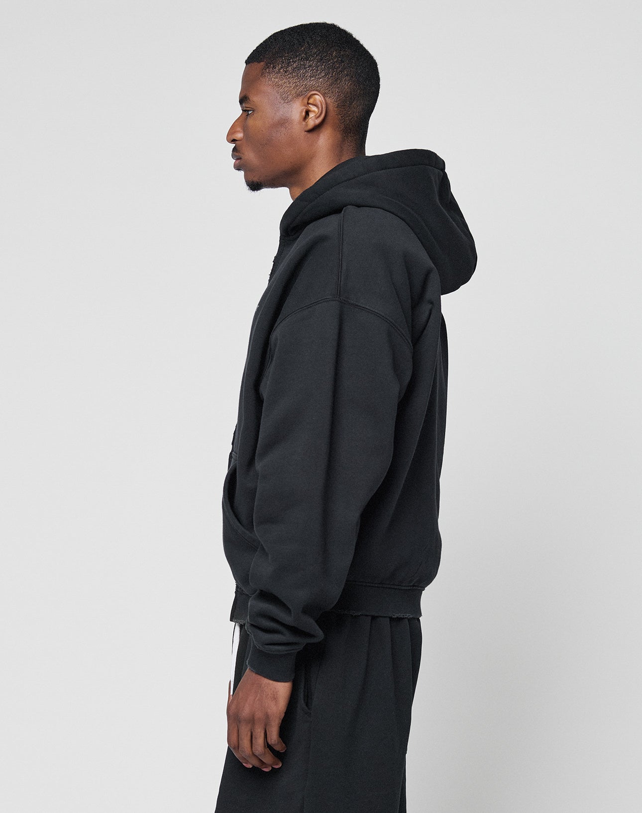 A man stands in profile against a plain background, wearing the LIVE FAST DIE YOUNG Basic Hooded Zip with subtle logo detail and black, slightly cropped pants, looking straight ahead with his arms relaxed by his sides.