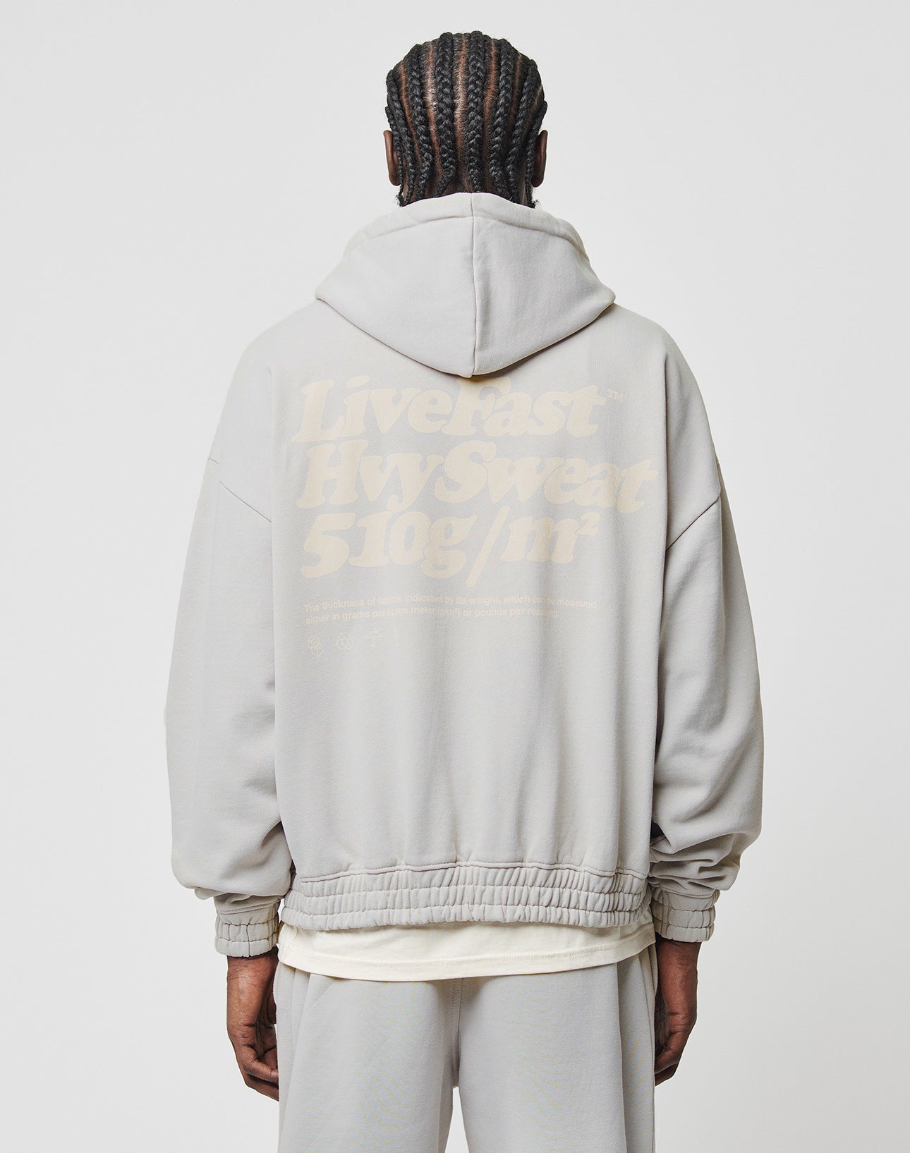 A person with braided hair, seen from behind, wears the LIVE FAST DIE YOUNG Basic 510 Hooded Zip—a light gray zip-up hoodie with a large faded “Livefast Hvysweat 510g/m²” print and stylish topstitching for an oversize fit.