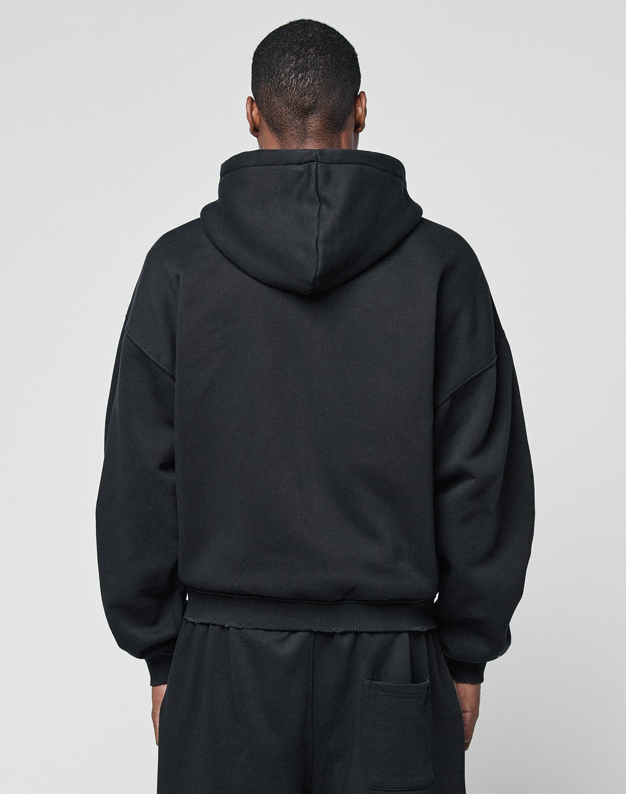 A person in a LIVE FAST DIE YOUNG Basic Hooded Zip and black pants stands facing away from the camera against a plain light gray background.