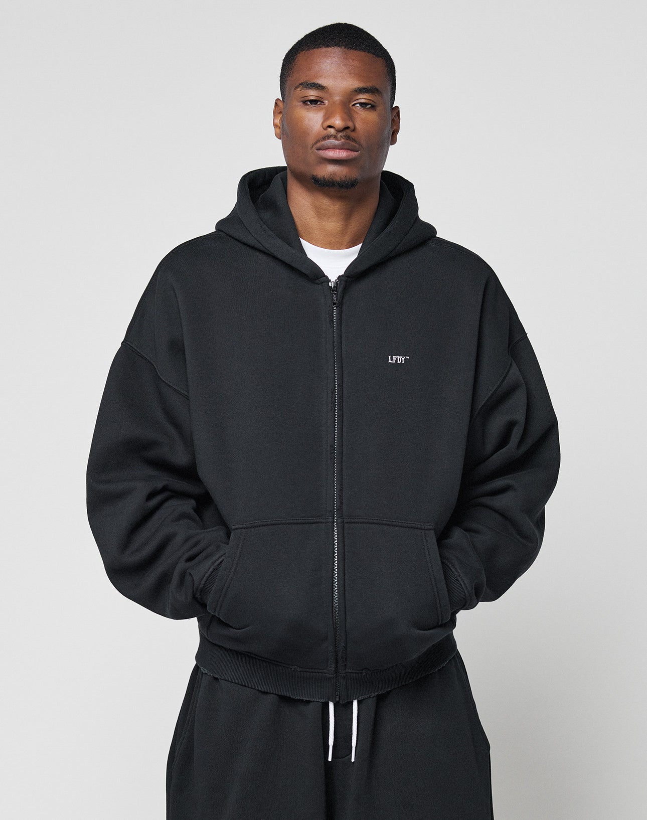 A man stands against a plain background, wearing the LIVE FAST DIE YOUNG Basic Hooded Zip in black with a small white logo print on the left chest. He has his hands in the pockets and displays a neutral expression.