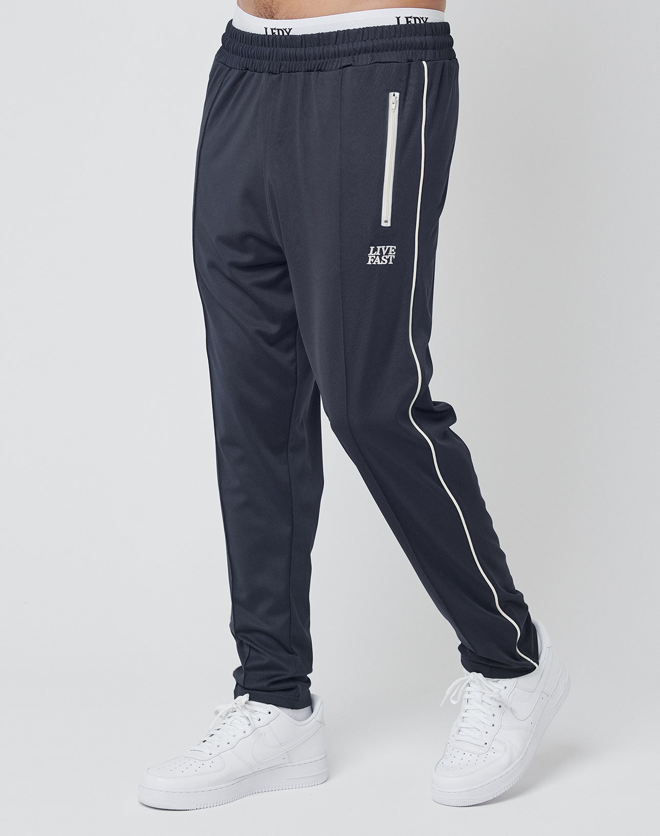A person wearing black Live Fast Trackjogger by LIVE FAST DIE YOUNG, featuring white piping, logo stitching, and LIFE ALES text on the left thigh, pairs it with white sneakers against a plain light background.
