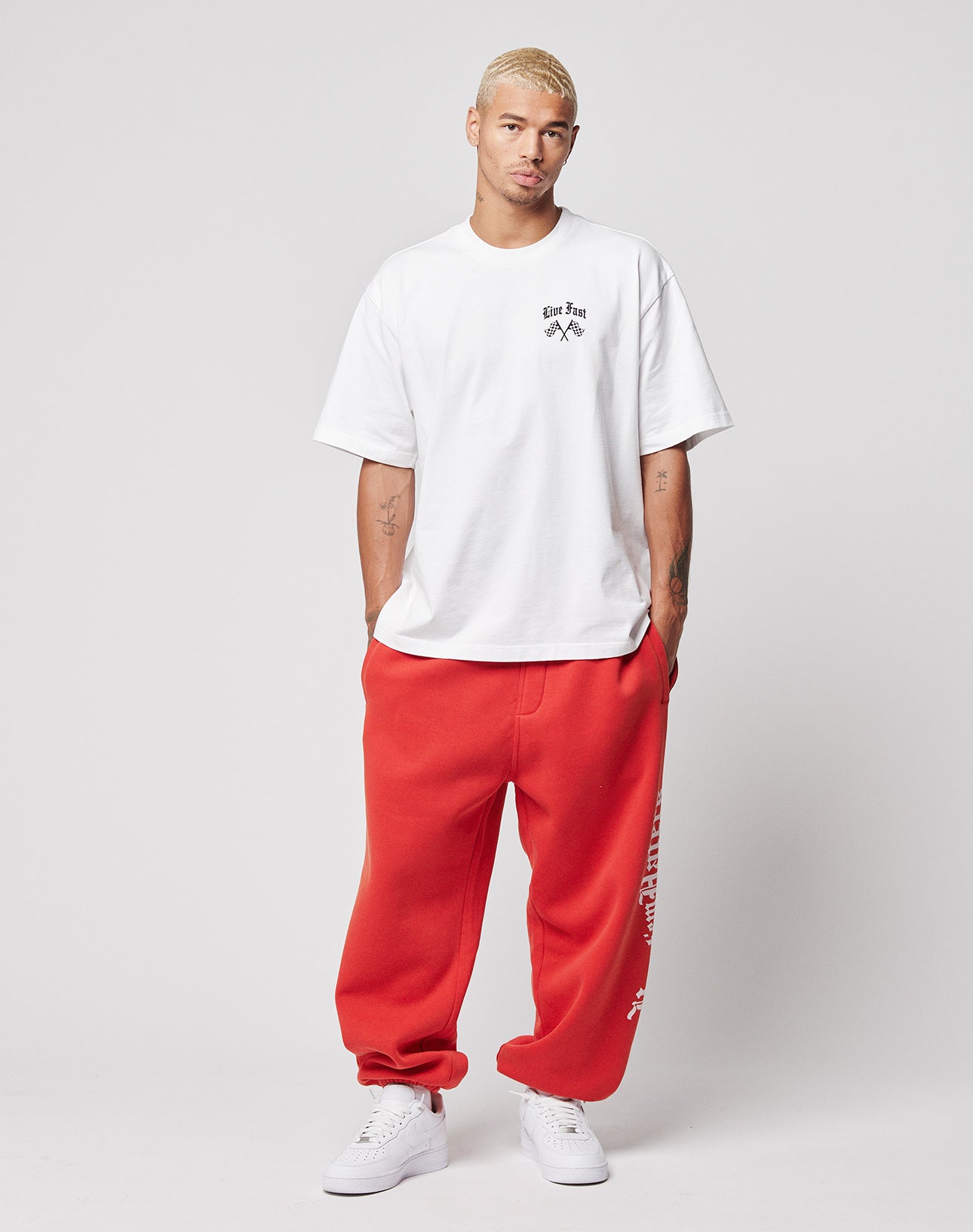 A man with short blond hair stands against a plain light background wearing a white graphic tee, white sneakers, and LIVE FAST DIE YOUNGās LFDY Haters Joggerāwide fit red sweatpants with an elastic waistband and white text on one leg.