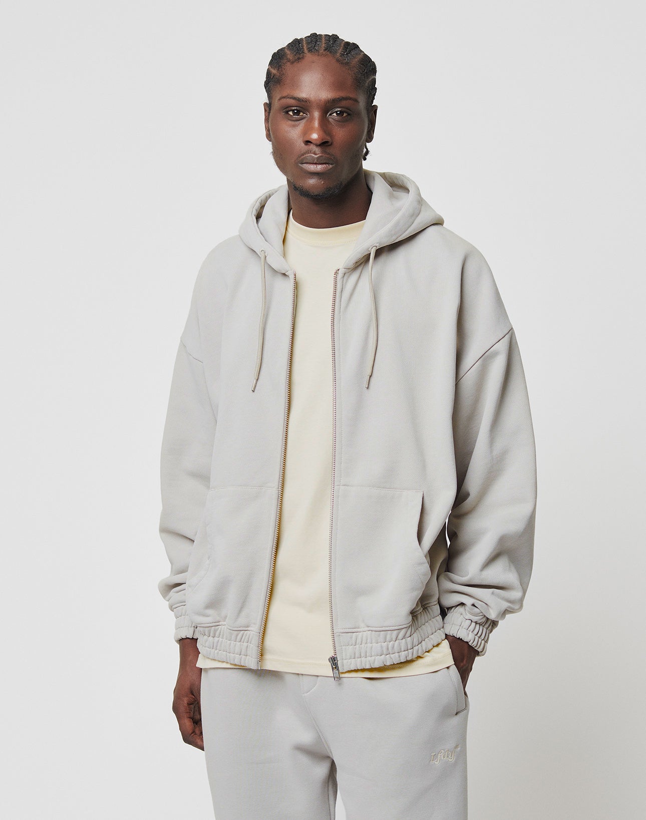 A man with braided hair wears the LIVE FAST DIE YOUNG Basic 510 Hooded Zip, an oversize light gray hoodie with backprint topstitching, over a beige T-shirt and matching gray pants, standing against a plain white background.