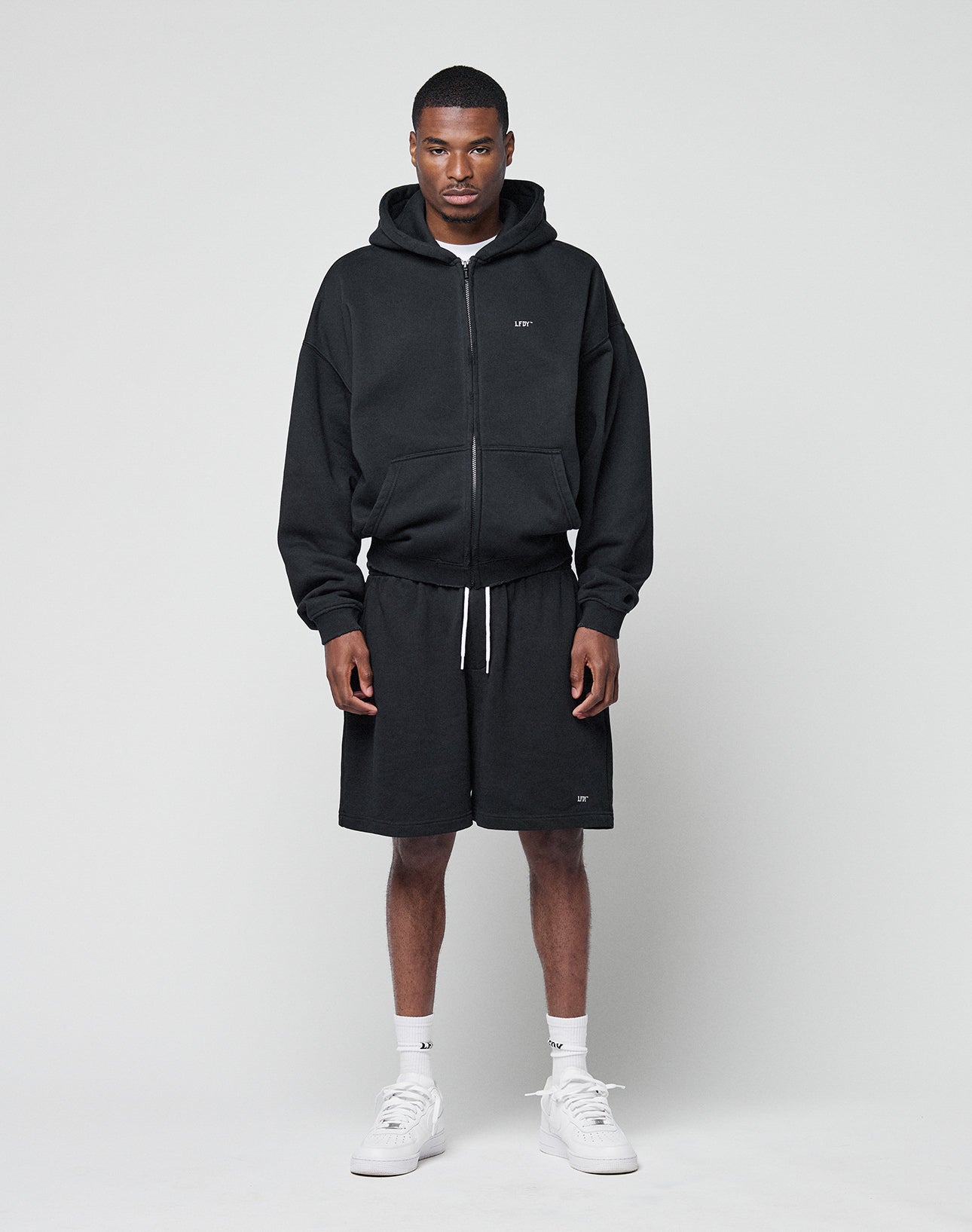 A man faces forward wearing the LIVE FAST DIE YOUNG Basic Hooded Zip in black with a logo print, paired with matching black shorts, white socks, and white sneakers against a plain light gray background.