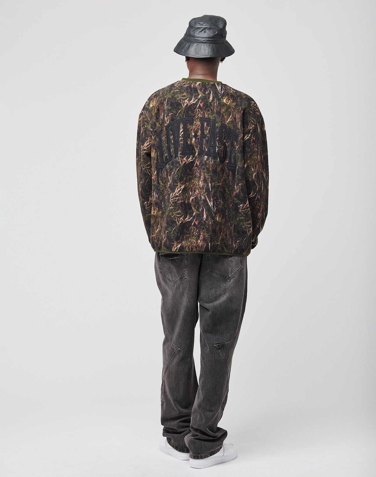 A person faces away in an oversize street style, wearing the LIVE FAST DIE YOUNG LFDY Fleece Longsleeve with bold text, a black bucket hat, loose dark jeans, and white sneakers against a plain light background.