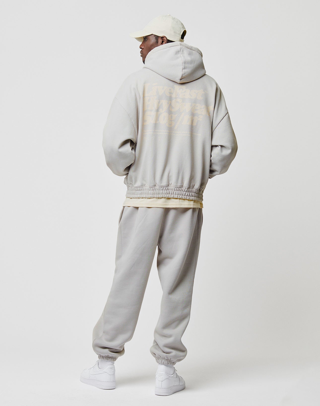 A person wearing the LIVE FAST DIE YOUNG Basic 510 Hooded Zip in light beige with backprint topstitching and matching sweatpants, plus a white cap and sneakers, stands facing away from the camera against a plain white background.