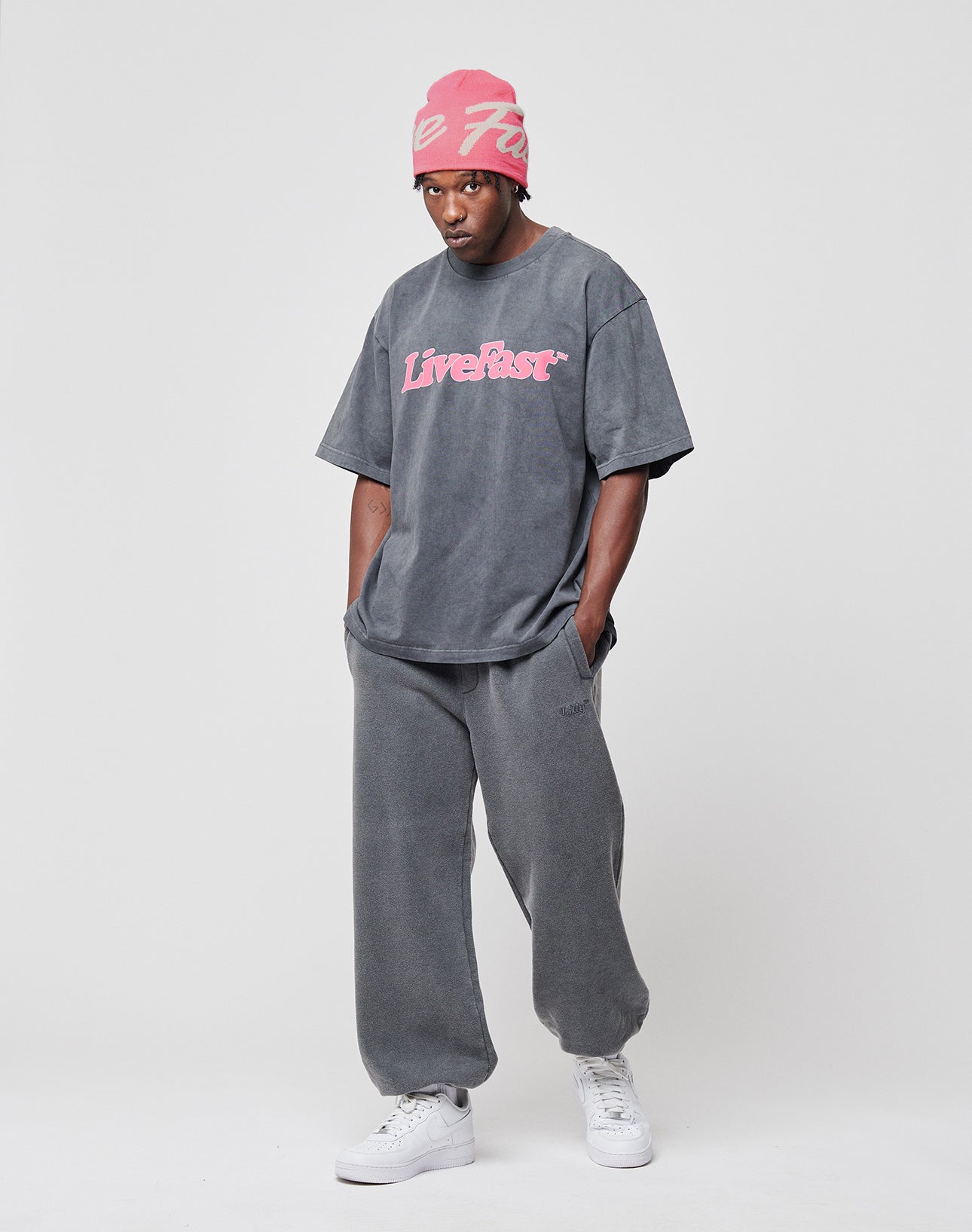 A man stands against a plain background wearing LIVE FAST DIE YOUNGs Loose Jogger in gray with elastik-bund and a loose fit gray Live Fast T-shirt, paired with a pink beanie and white sneakers. His hands are in his pockets, looking serious.
