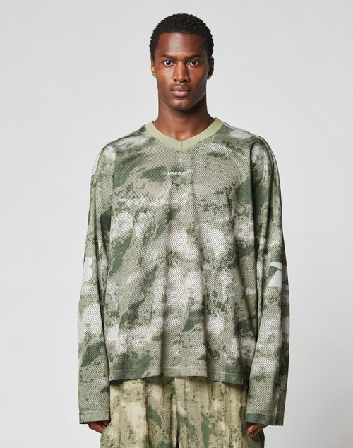 A man faces forward against a plain light background, wearing the LIVE FAST DIE YOUNG Camo Mesh Jersey—an oversized, long-sleeved green and beige camo shirt with a reflective print, number 23 on the sleeves—and light pants.