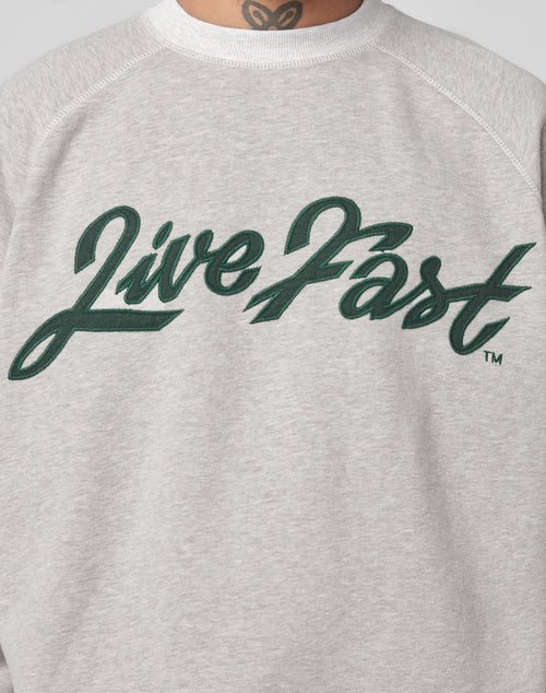 A person wears the LFDY Script Sweater by LIVE FAST DIE YOUNG, an oversize fit light gray sweatshirt with raglan sleeves and Live Fast in large green cursive on the front. A neck tattoo is visible above the collar.