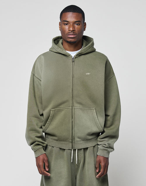A man faces forward wearing an olive green Basic Hooded Zip and matching sweatpants from LIVE FAST DIE YOUNG, featuring a subtle faded look and a small white LIFY logo on the chest. The background is plain light gray.