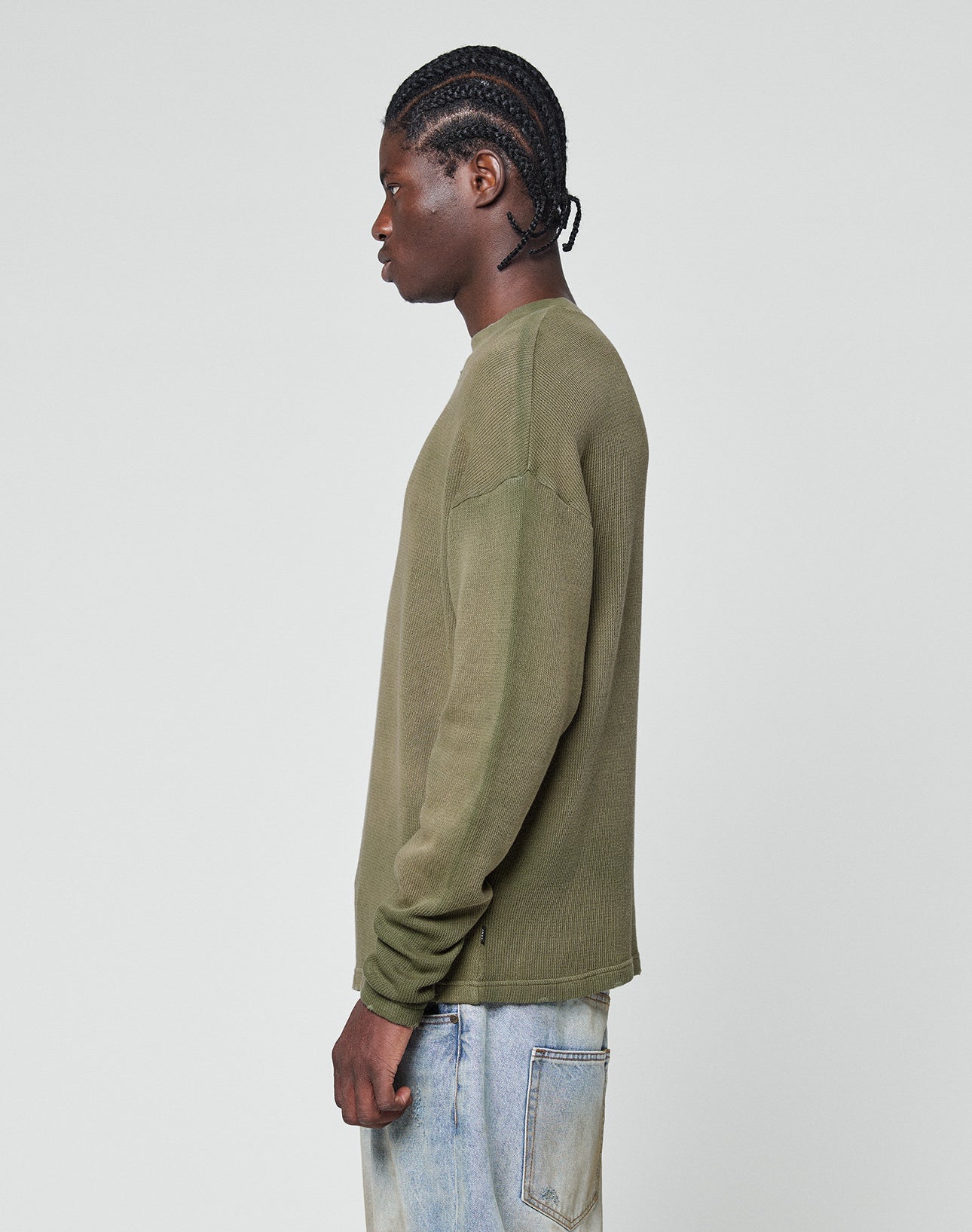 A person with braided hair stands in profile against a plain light background, wearing the LIVE FAST DIE YOUNG LFDY Rib Knit Longsleeve in olive green and light blue jeans with individuellem Wasch-Effekt and visible back pocket stitching.