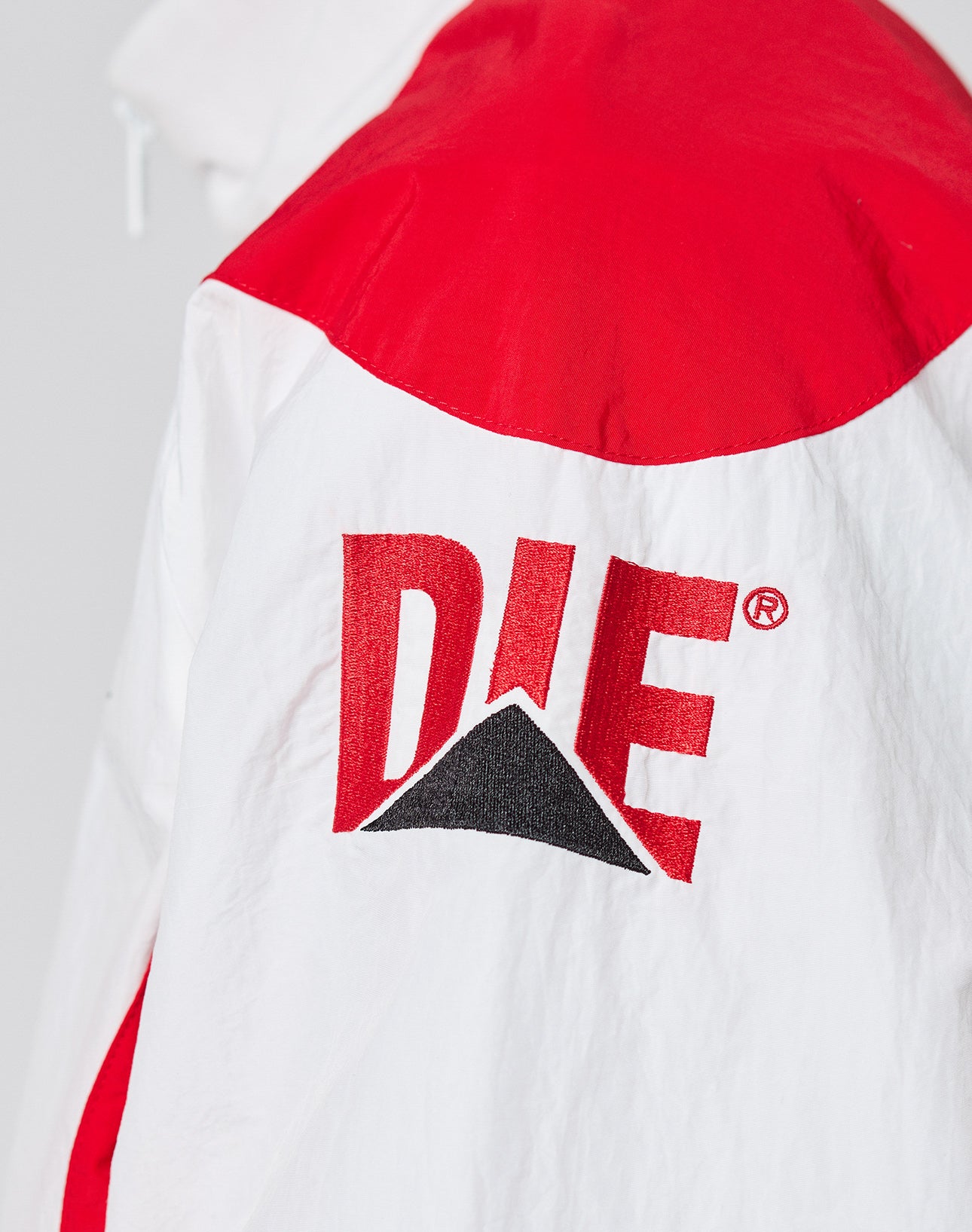 A close-up of the LFDY Trackjacket 3000 by LIVE FAST DIE YOUNG shows a white jacket with red shoulder panel and bold red DIE text above a black triangle on the back. Regular Fit with Branded ReiĆverschluss-Puller adds stylish detail.