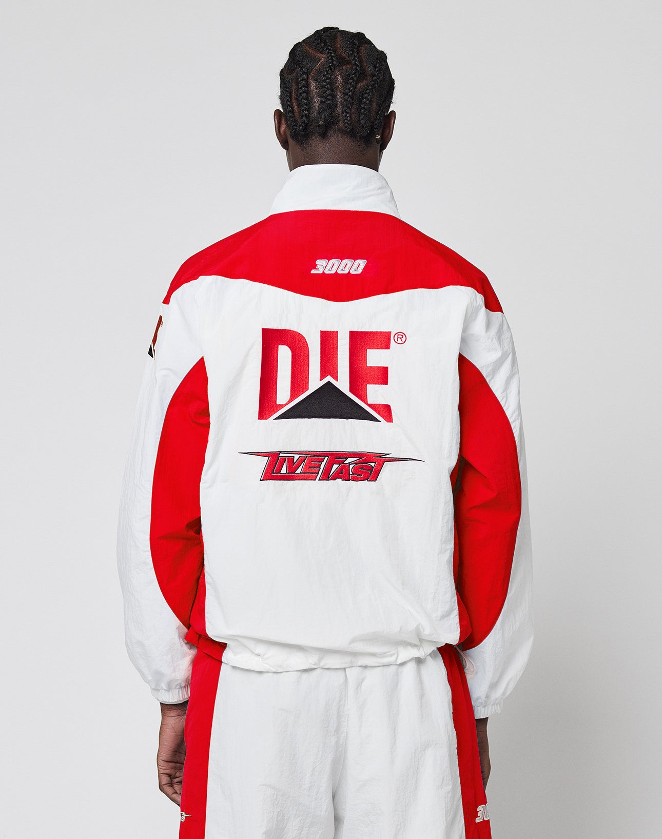 A person with braided hair, seen from behind, wears the LIVE FAST DIE YOUNG LFDY Trackjacket 3000āa regular fit red and white jacket with bold āDIE,ā āLIVE FAST,ā graphic designs, multiple logos, and the number 3000 near the collar.