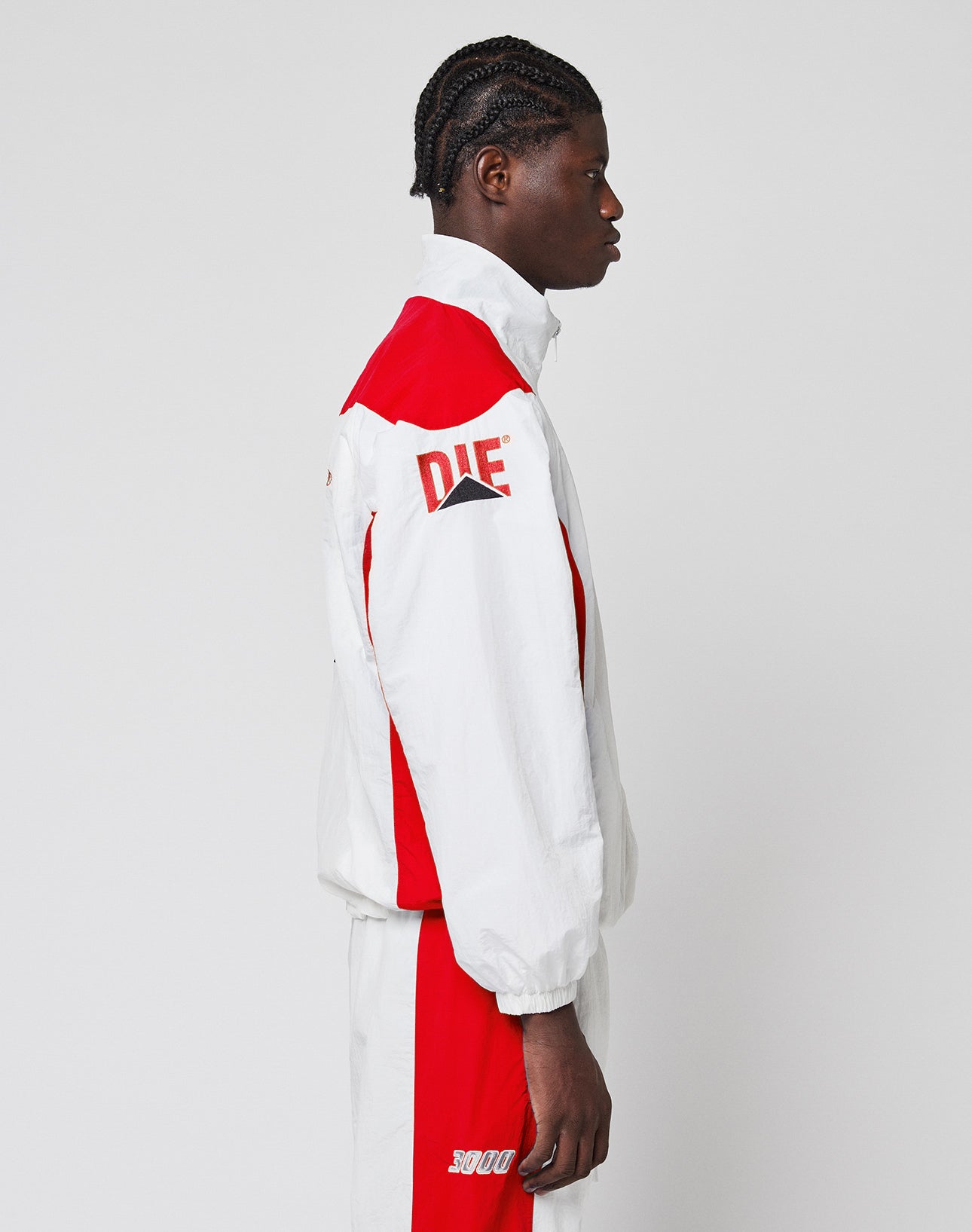 A person with braided hair stands in profile, wearing the LIVE FAST DIE YOUNG LFDY Trackjacket 3000āa regular-fit, color-block tracksuit with DIE on the sleeve, 5000 on the shorts, and an elastischer Kordelzug.