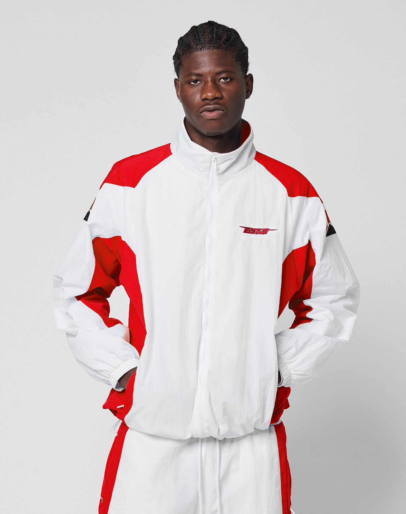 A man wears the LIVE FAST DIE YOUNG LFDY Trackjacket 3000 in regular fit white and red with branded zip puller, standing against a light gray background, hands in pockets, looking at the camera with a neutral expression.