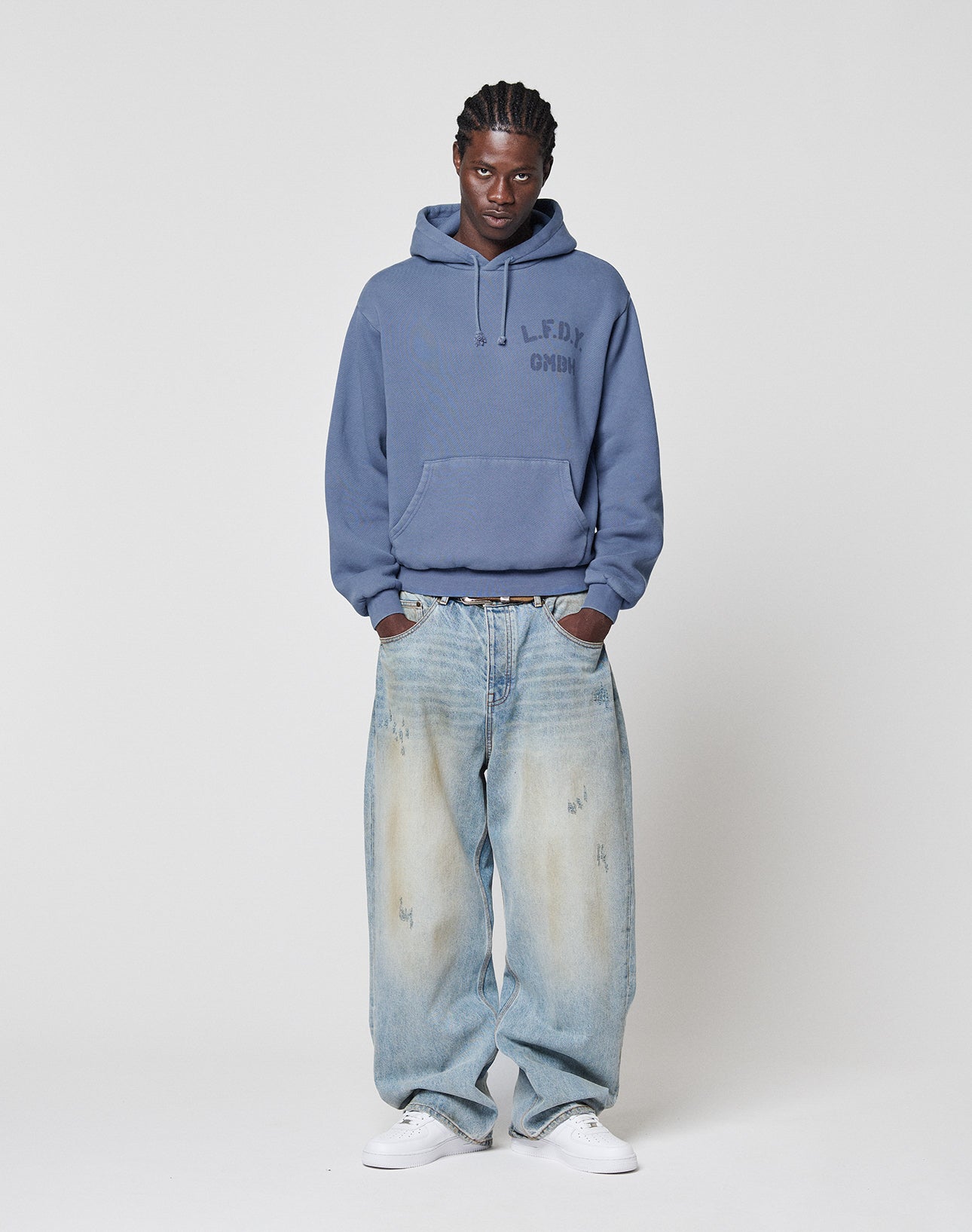 A person stands against a plain background wearing a blue LIVE FAST DIE YOUNG LFDY GmbH Hooded, loose-fitting faded jeans with Vintage Waschung, and white sneakers, hands in pockets with a neutral expression.