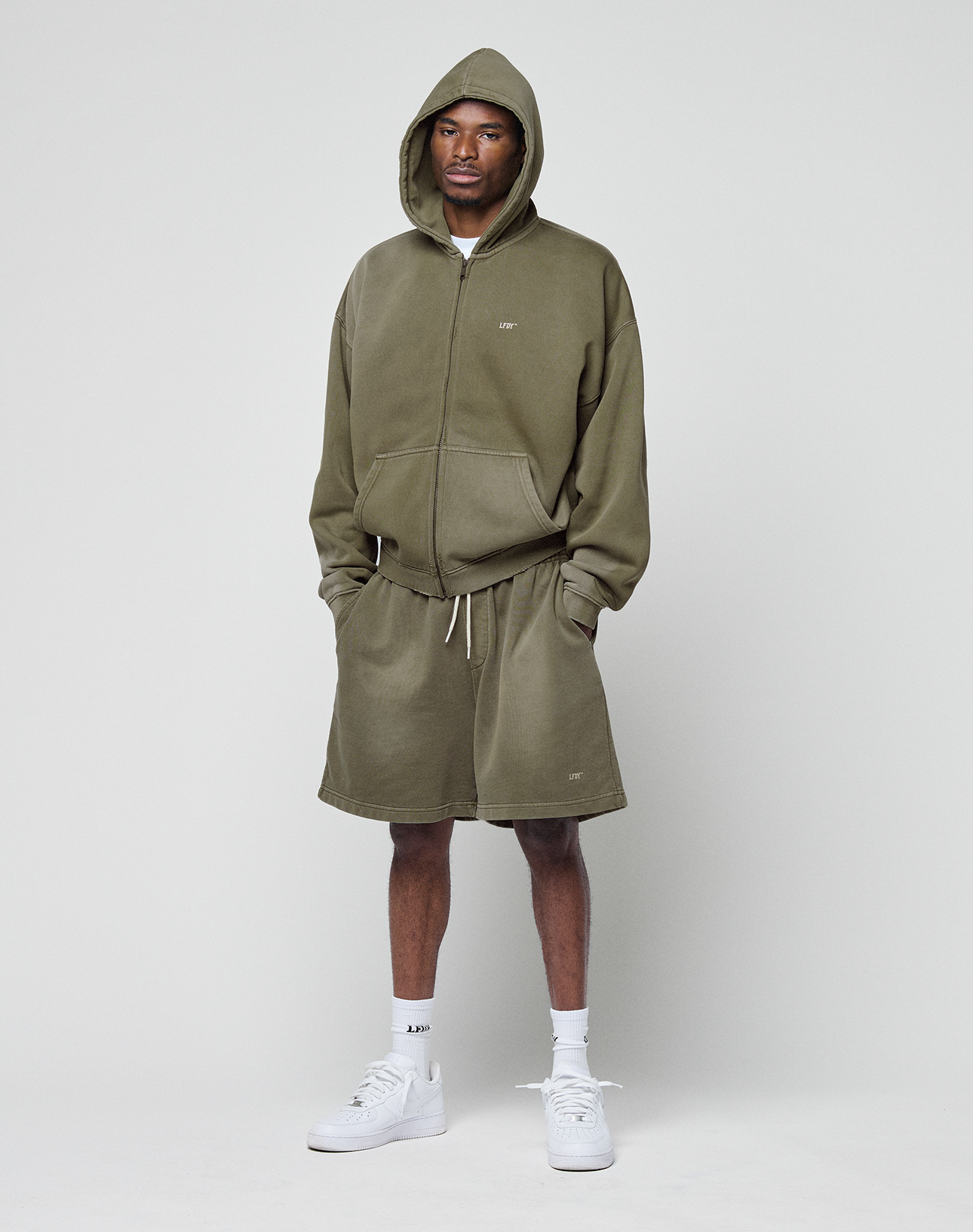 A man stands against a white background wearing the LIVE FAST DIE YOUNG Basic Hooded Zip in olive green with a subtle logo and oversize fit, matching shorts, white socks, and sneakers. Hands in pockets, he looks directly at the camera.