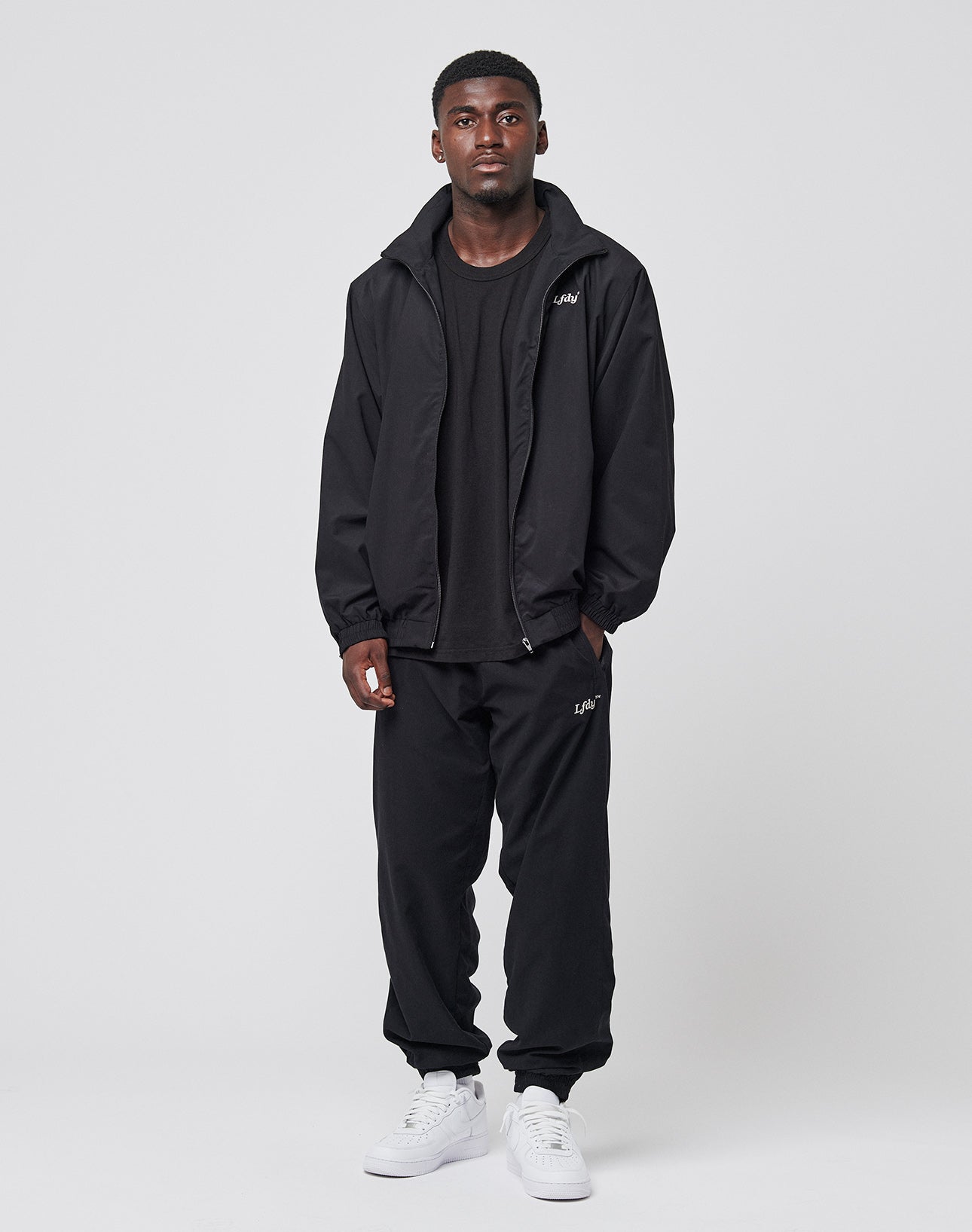 A man stands against a plain light gray background, wearing the LIVE FAST DIE YOUNG LFDY Trackjacket with elastic cuffs, a black t-shirt, black jogger pants with side pockets, and white sneakers. His expression is neutral and his arms are relaxed at his sides.