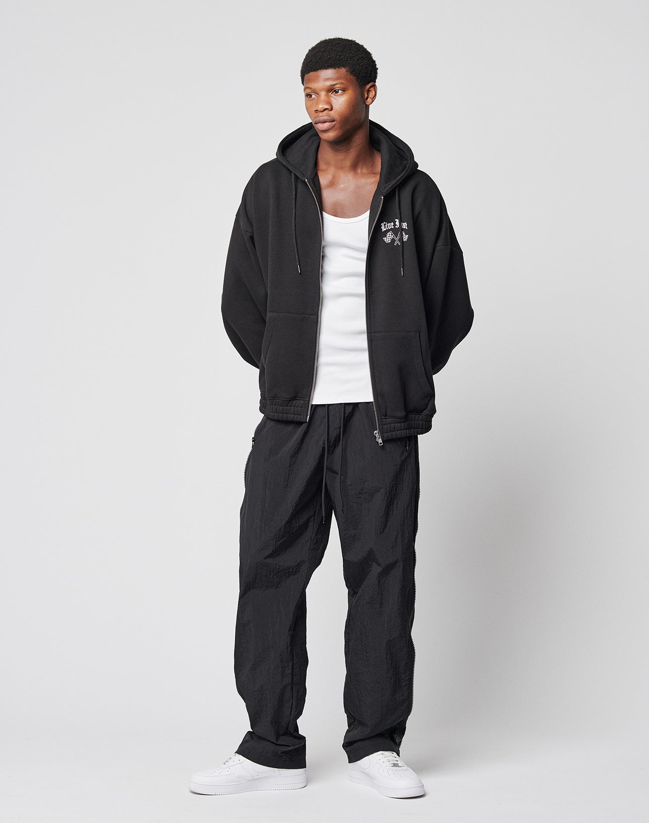 A man stands against a plain background wearing LFDY Zip Tape Track Pants, a black zip-up hoodie over a white tank top, and white sneakers. His hands are in his pockets and hes looking slightly to the side.