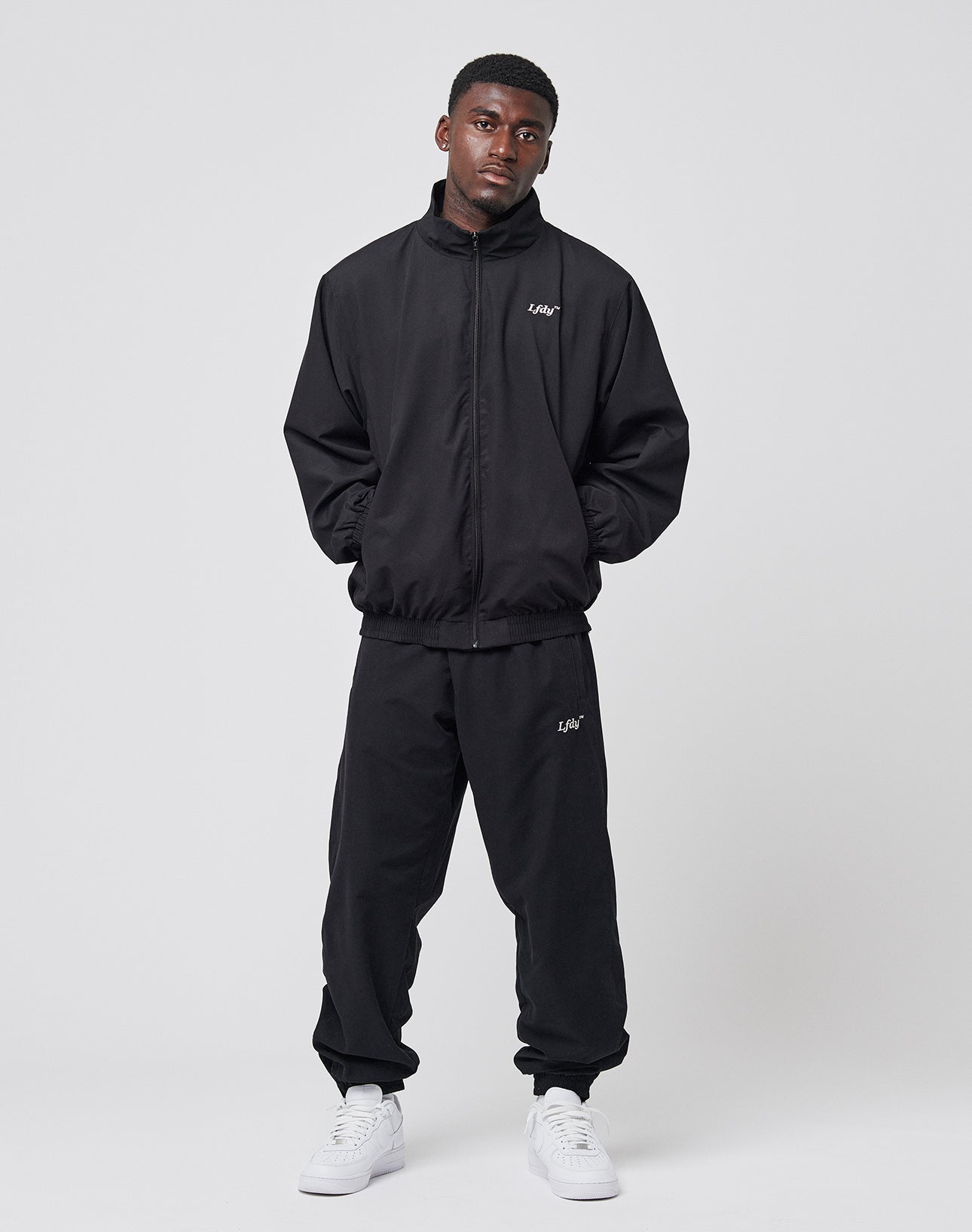 A man stands against a plain light background wearing black LFDY Trackpants by LIVE FAST DIE YOUNG with Reißverschluss, white sneakers, hands in pockets, and looking slightly upward.