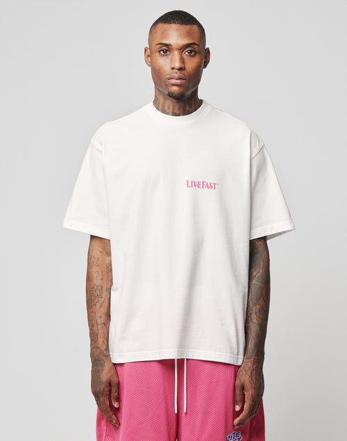 A tattooed man wears the LFDY Loyal Tee by LIVE FAST DIE YOUNG—an oversized white cotton T-shirt with pink LIVE FAST front print—paired with pink athletic shorts, standing against a plain gray background.
